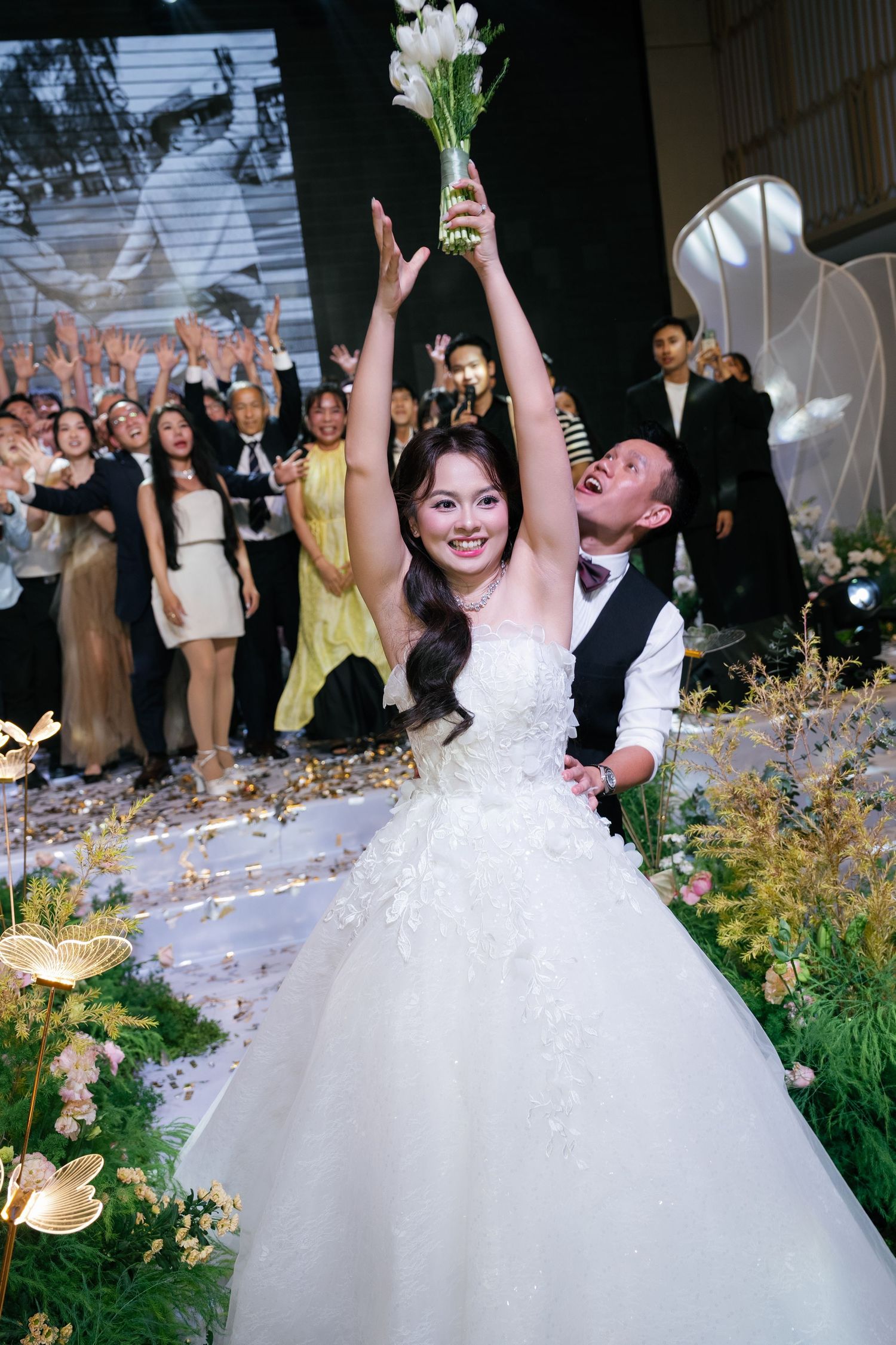 A joyful bride in a white floral gown raises her bouquet while groom stands behind her at wedding.
