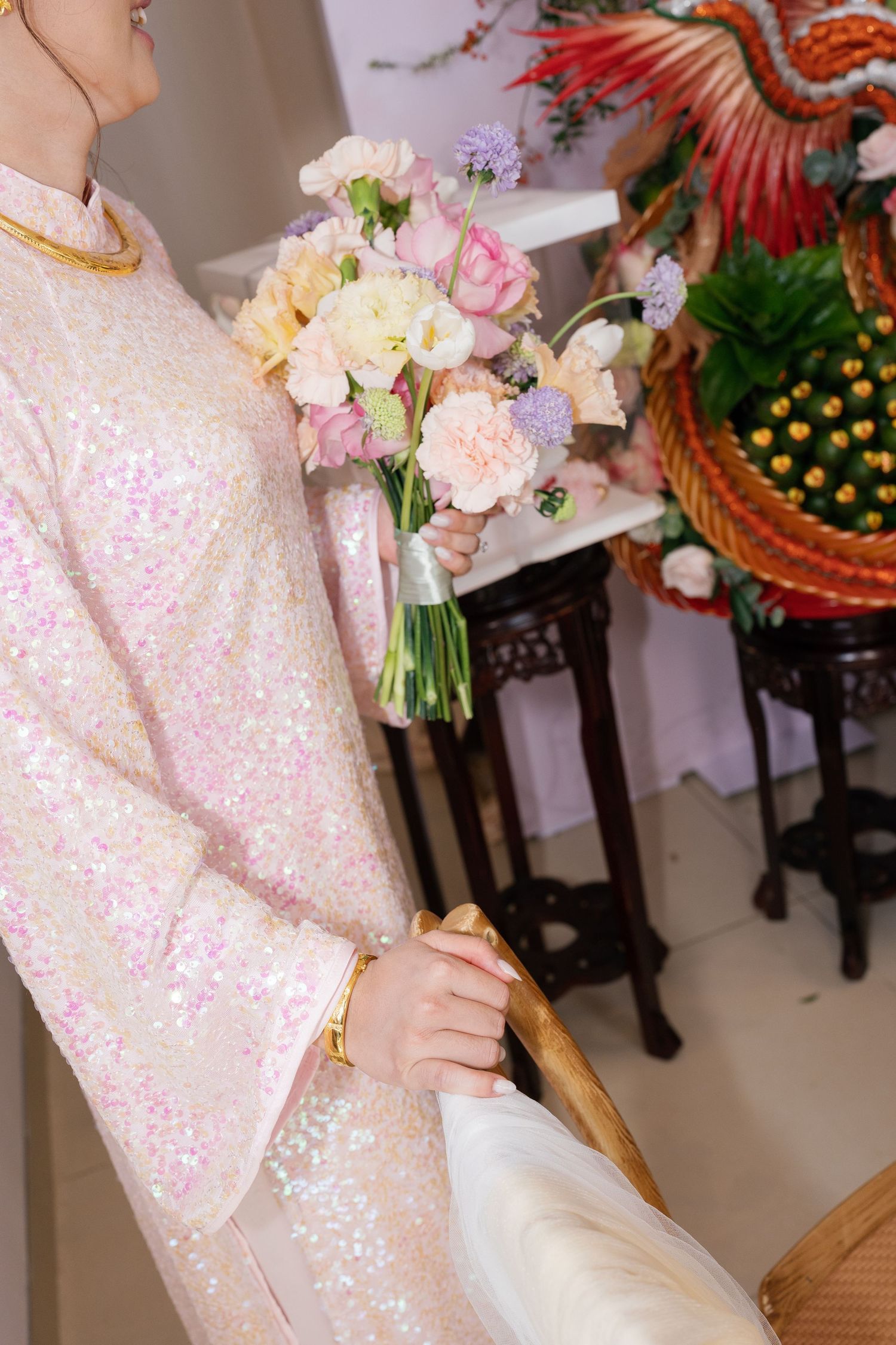Bride in pink sequin ao dai holding pastel wedding bouquet with roses and wildflowers at traditional Vietnamese ceremony.