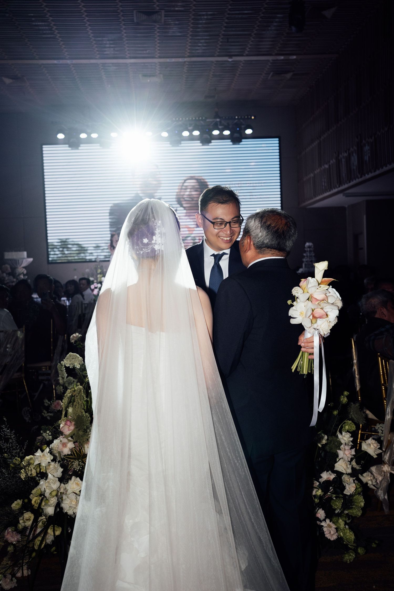Asian bride in white gown and veil walks down aisle with father toward smiling groom at elegant wedding ceremony.