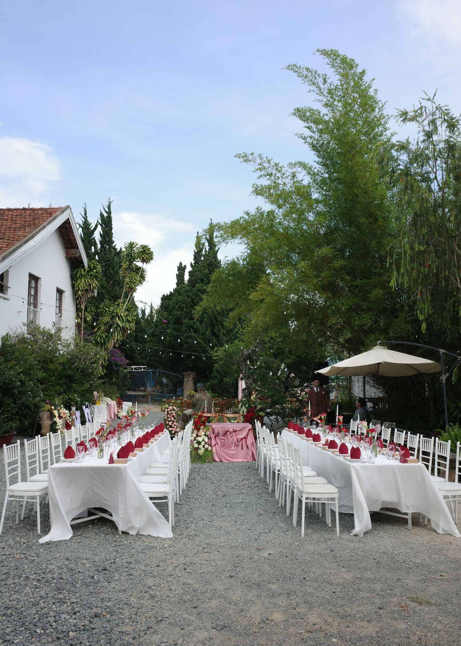 Elegant outdoor wedding reception with long white-draped tables, red floral accents, and white chairs set in a lush garden.