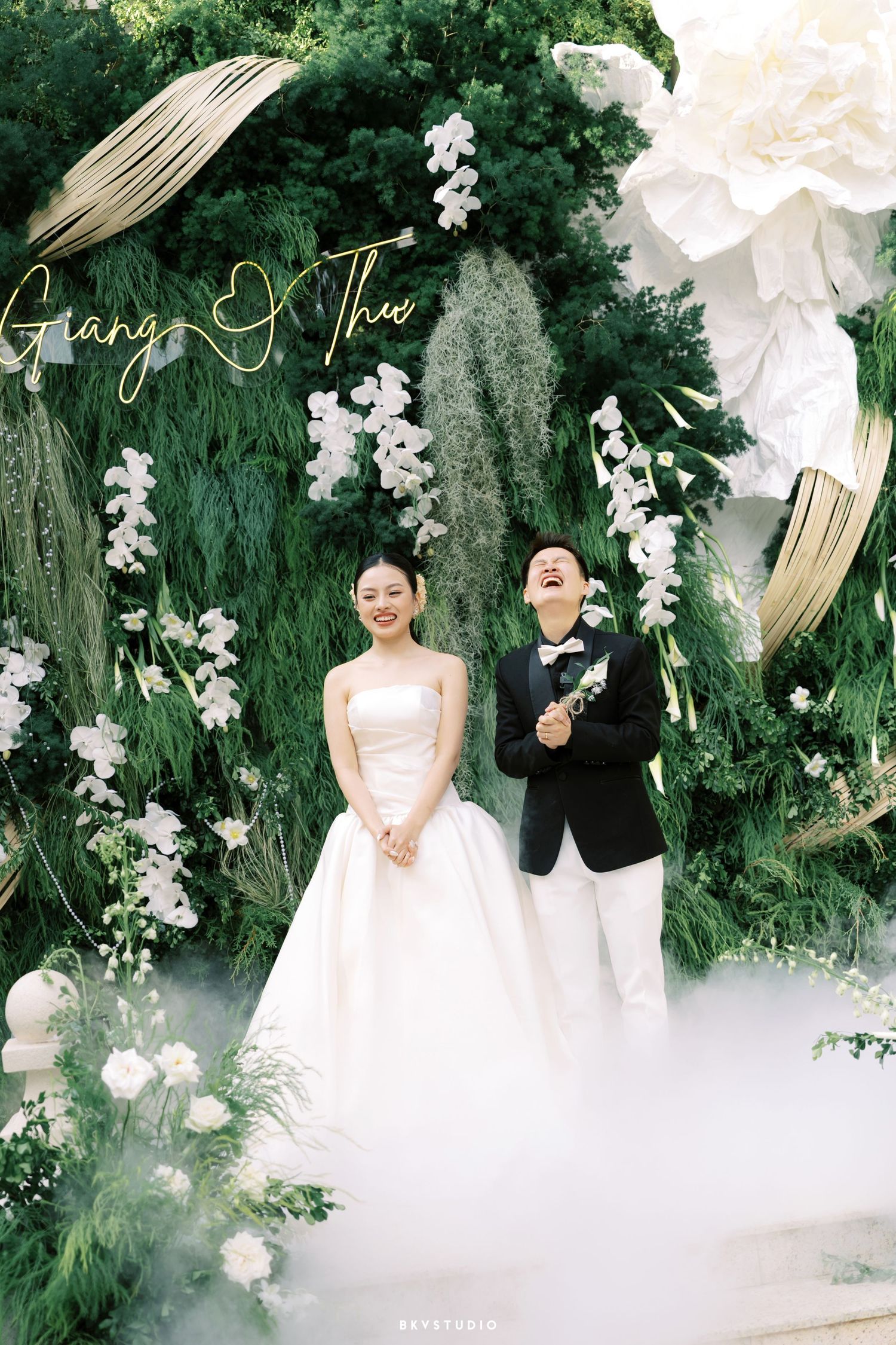 Laughing bride and groom pose before lush green floral backdrop with neon name sign.