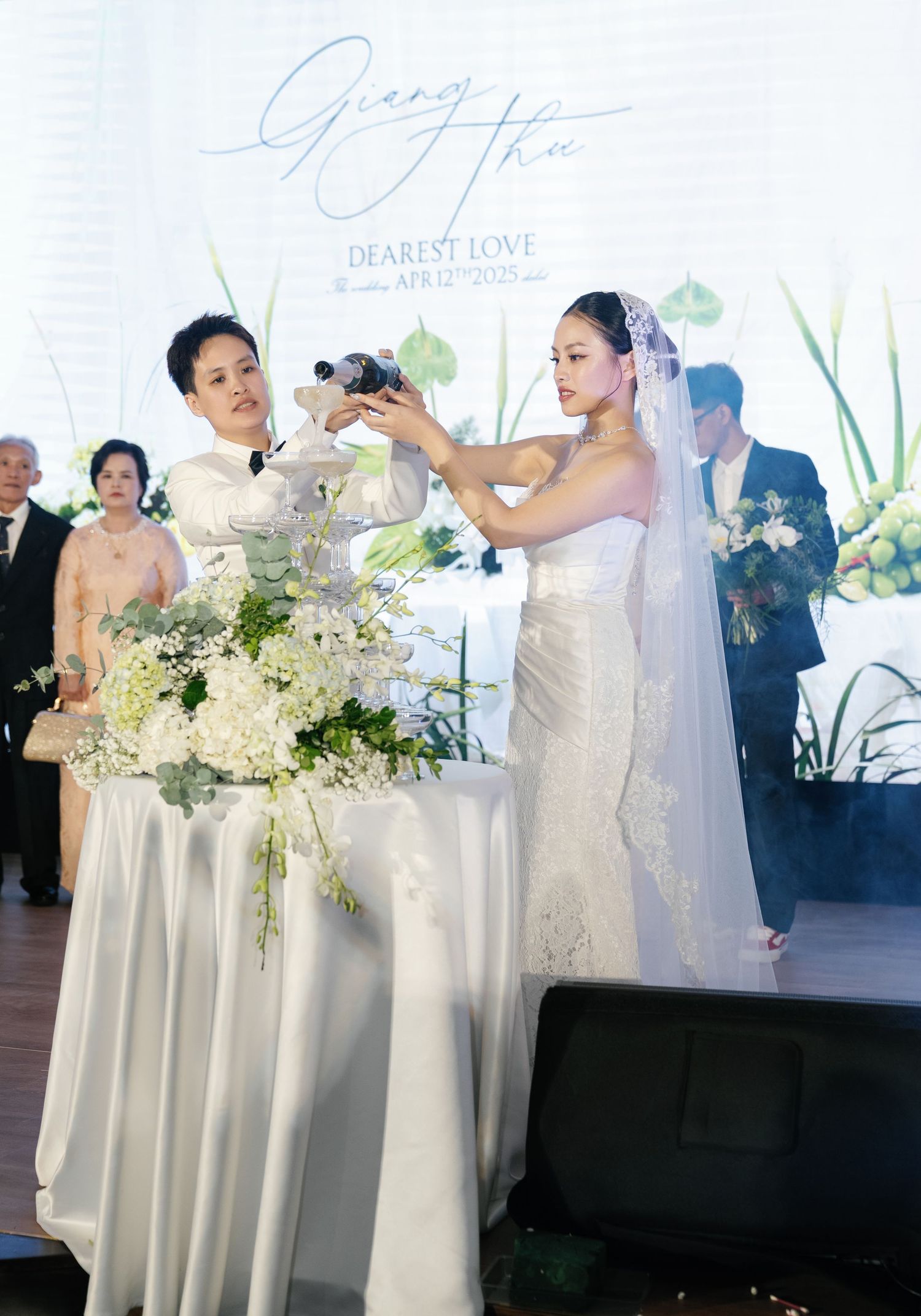 Bride in lace wedding gown and veil participates in tea ceremony with family member indoors.