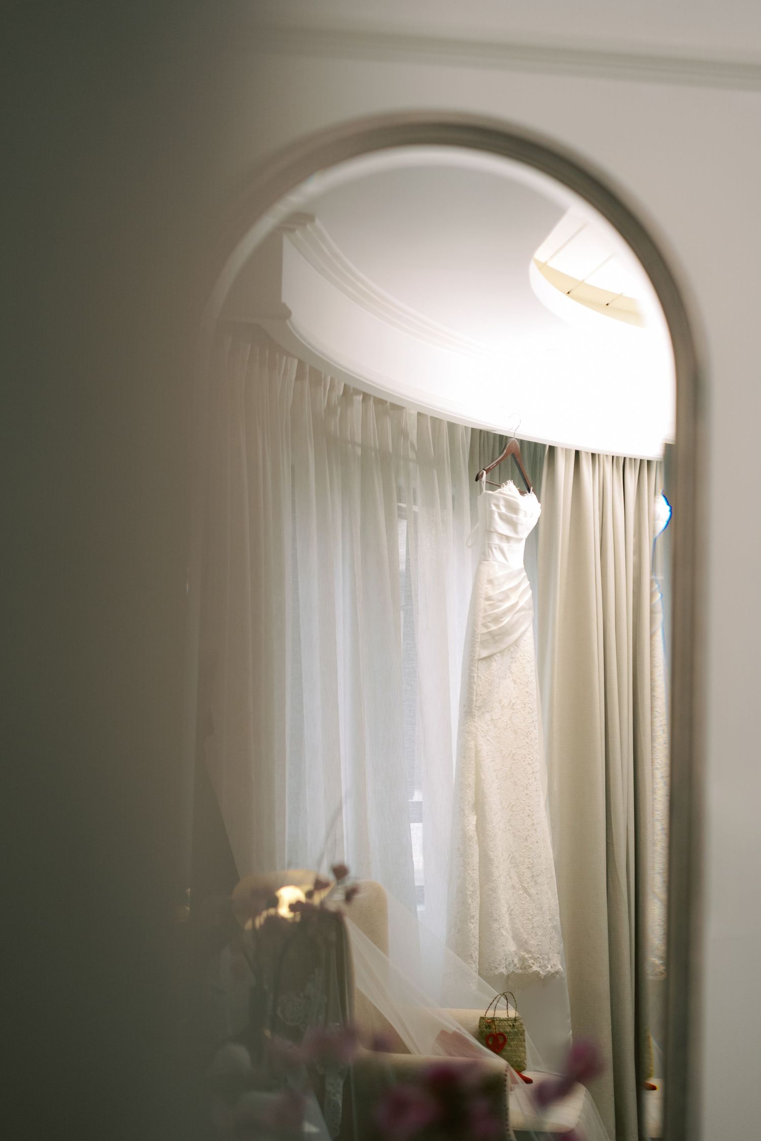 Wedding dress hanging by a window reflected in an arched mirror, soft natural light illuminating the bridal gown.