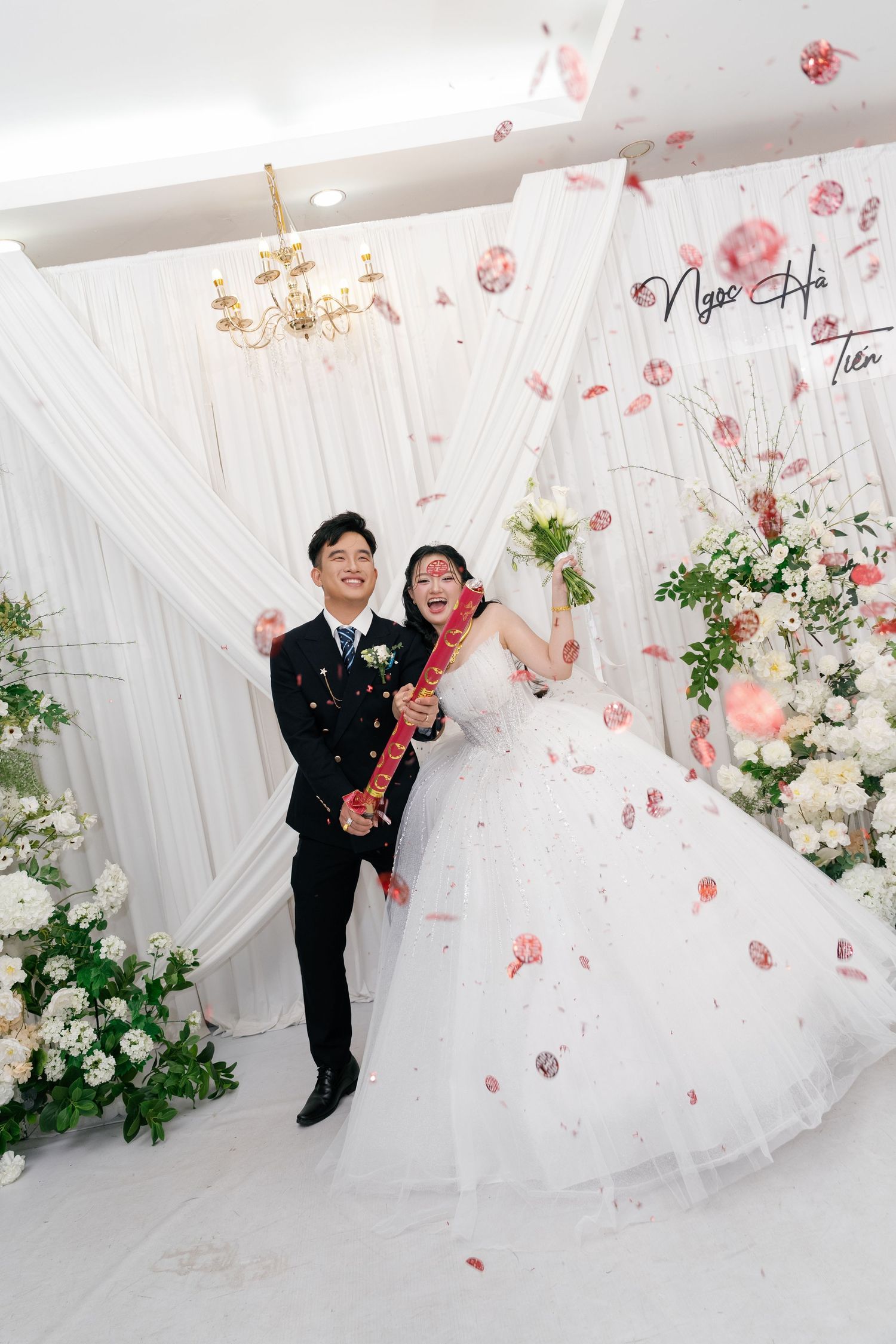 Joyful bride and groom celebrate with confetti poppers in front of a white floral wedding backdrop.