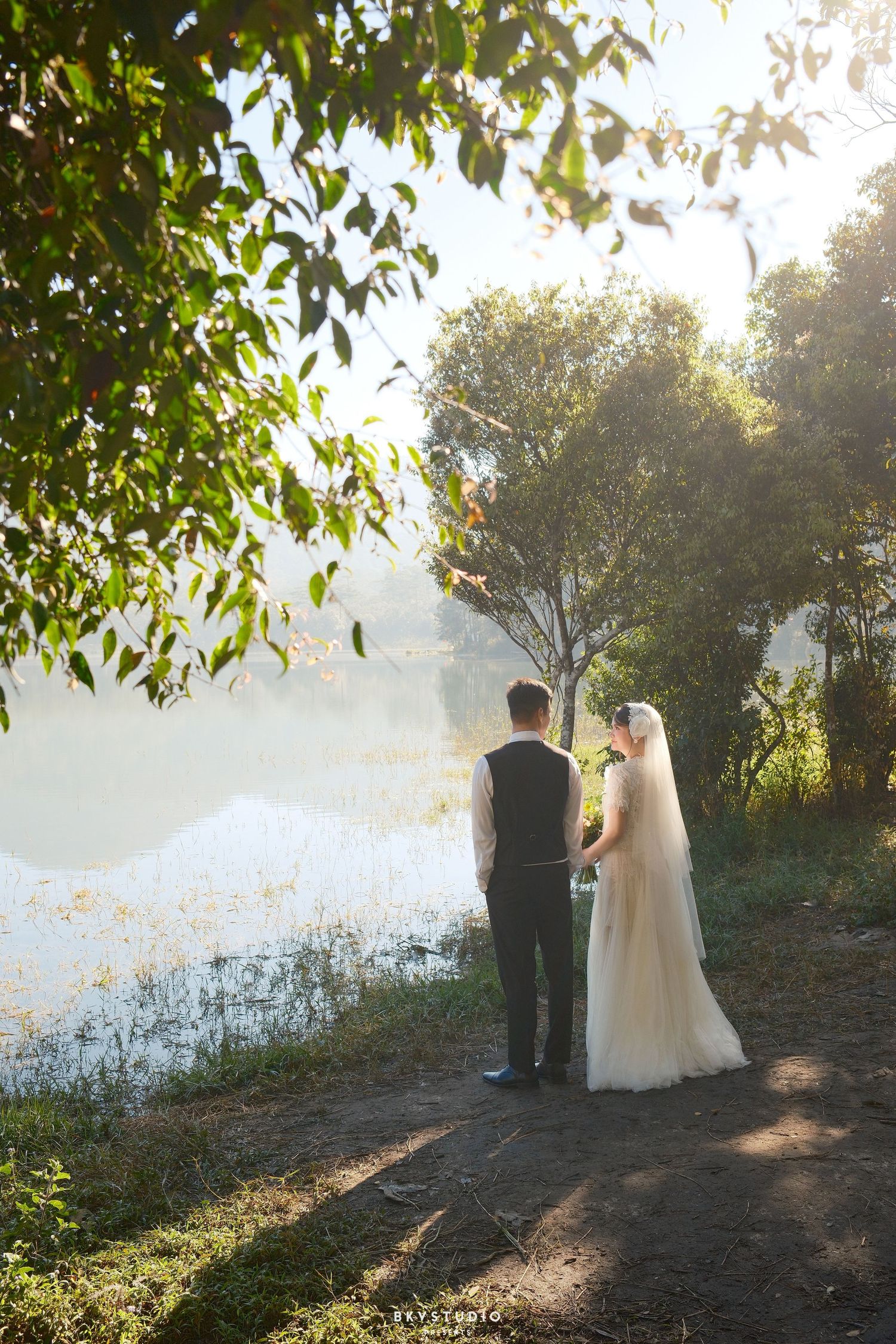 Bride and groom stand hand-in-hand by a misty sunlit lake surrounded by lush green trees.
