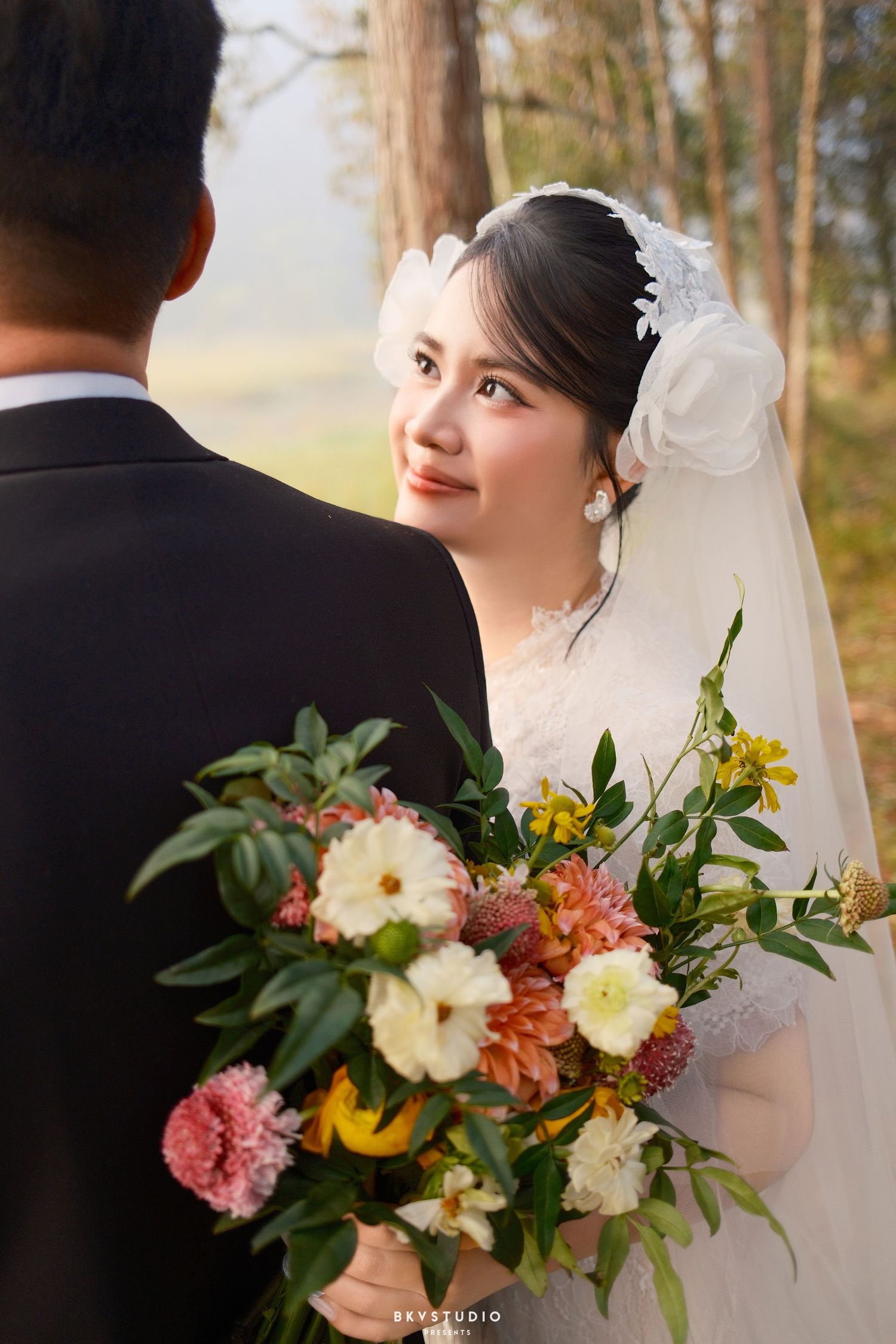 Bride in white lace gown and veil smiles at groom holding colorful bouquet outdoors.