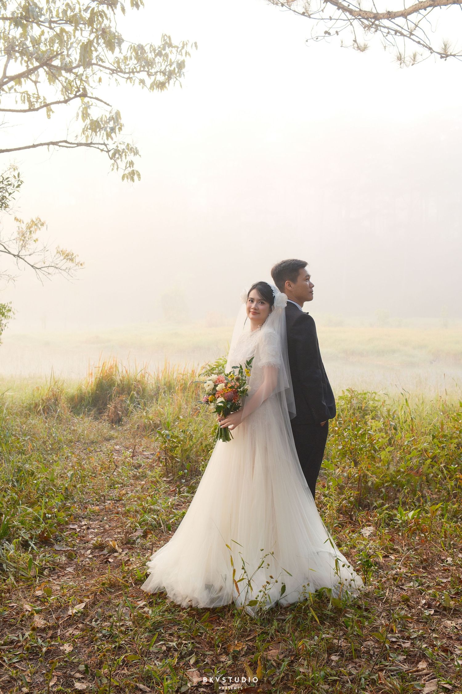 Bride and groom stand back-to-back in misty meadow, bride holding colorful bouquet in white gown.