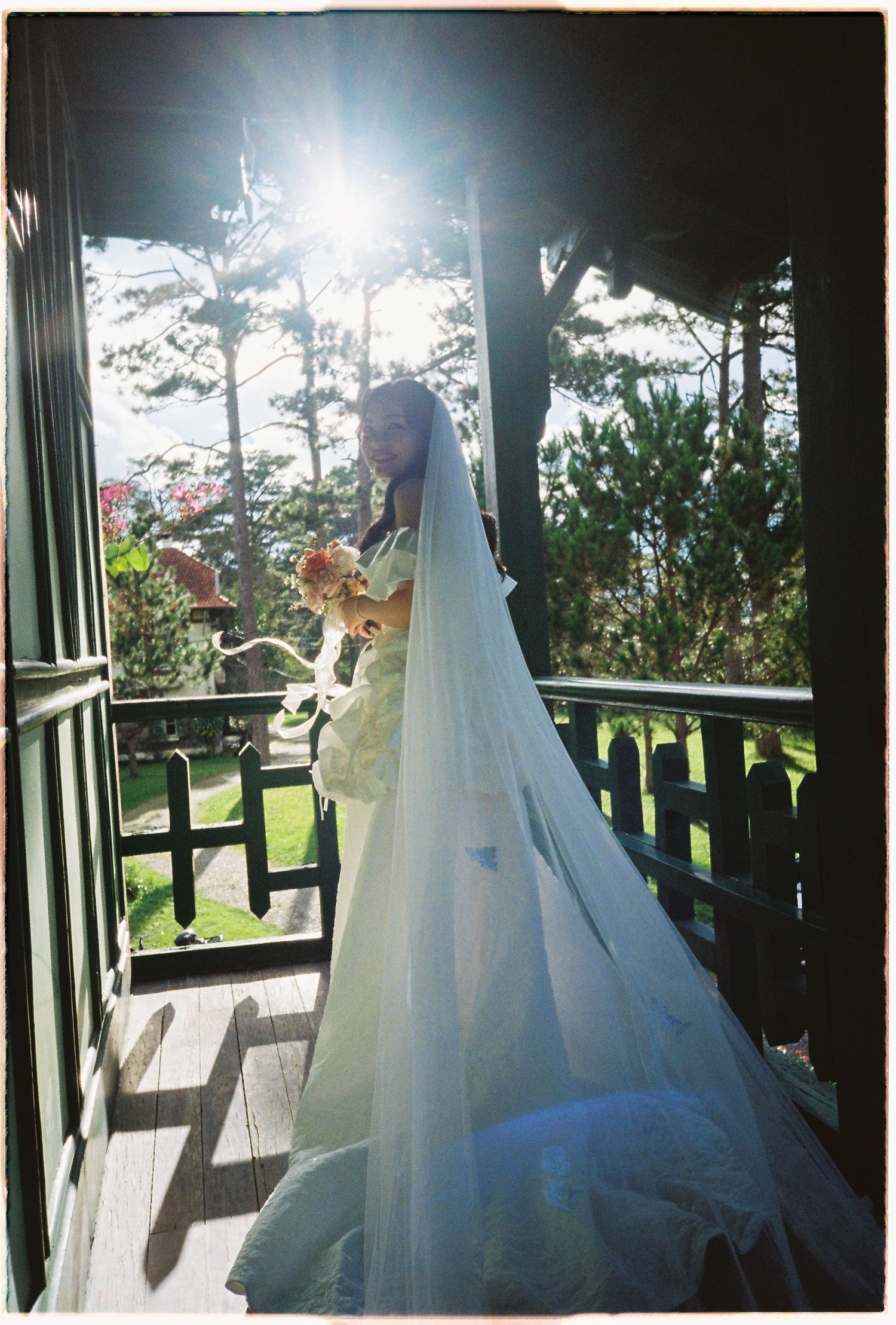 Bride in white wedding gown and long veil holding bouquet on sunlit wooden porch surrounded by lush green trees.