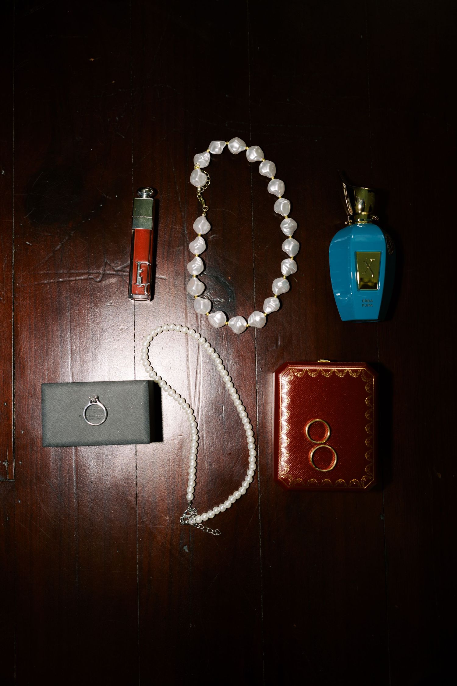 Pearl necklace, diamond ring, gold wedding bands, blue perfume bottle, and lipstick arranged on dark wood surface.