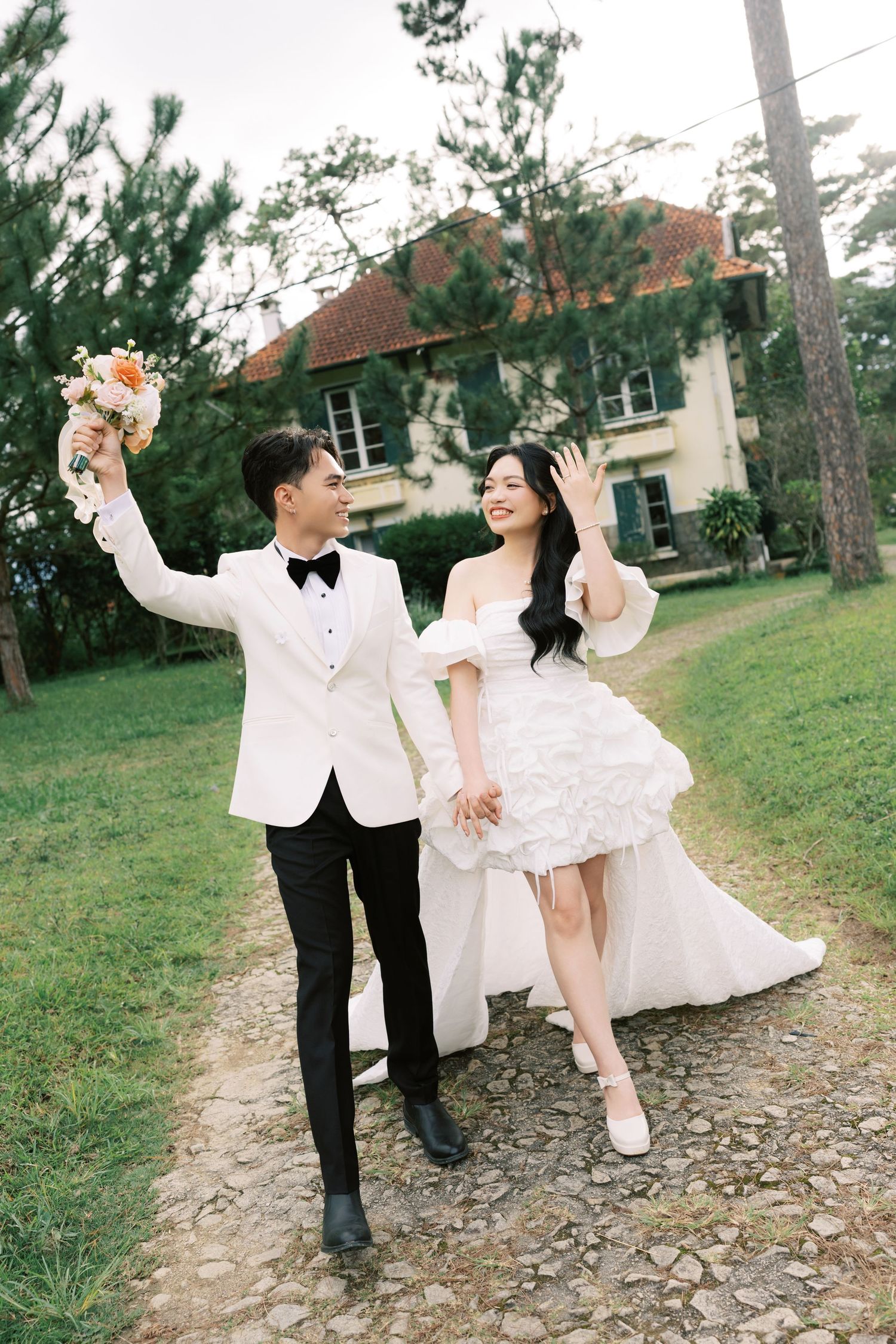 A joyful Asian couple in wedding attire walks hand-in-hand on a garden path, groom in white tuxedo, bride in ruffled gown.