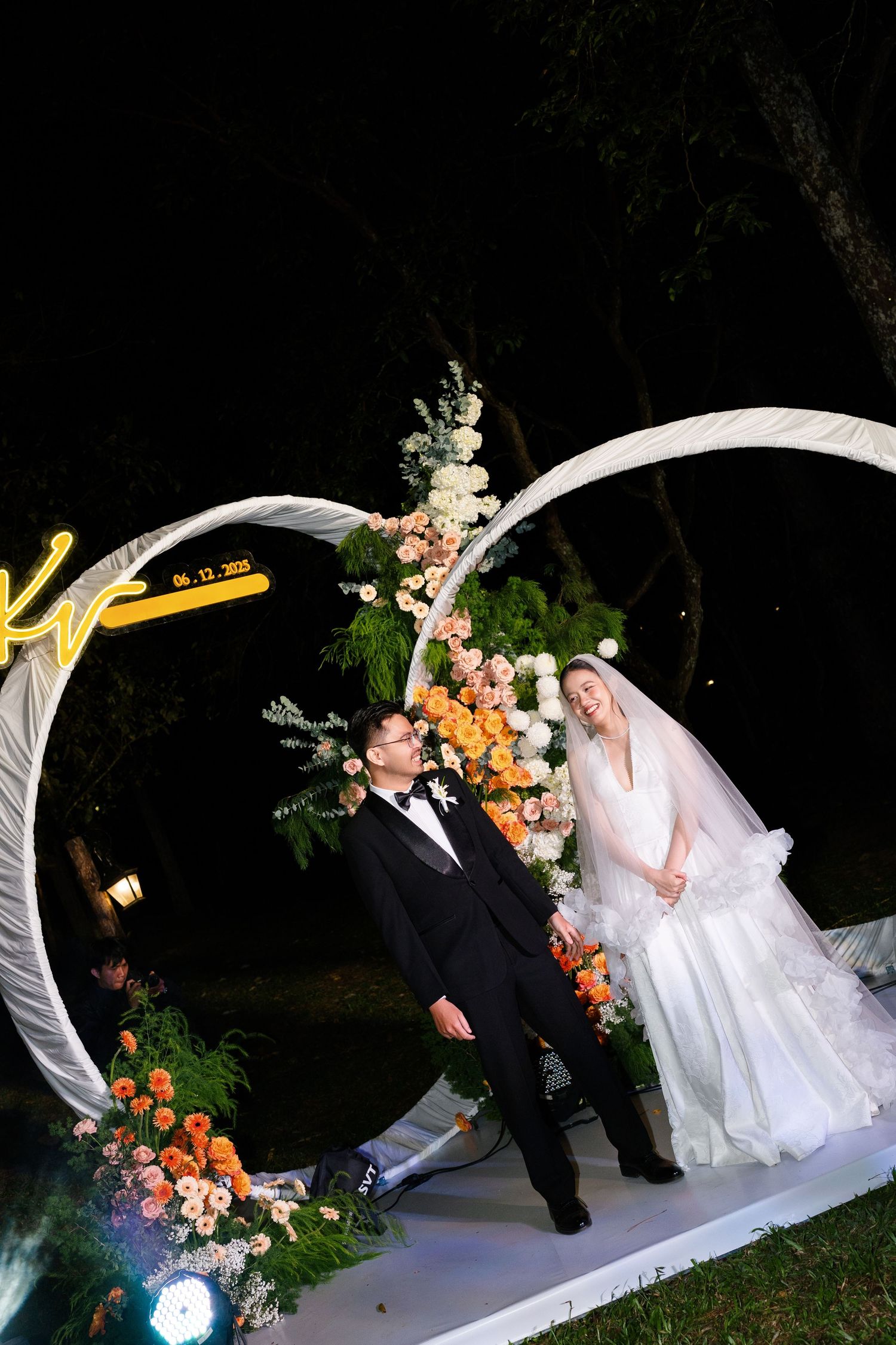 Bride in white gown and groom in tuxedo laugh together under floral arch at night wedding.