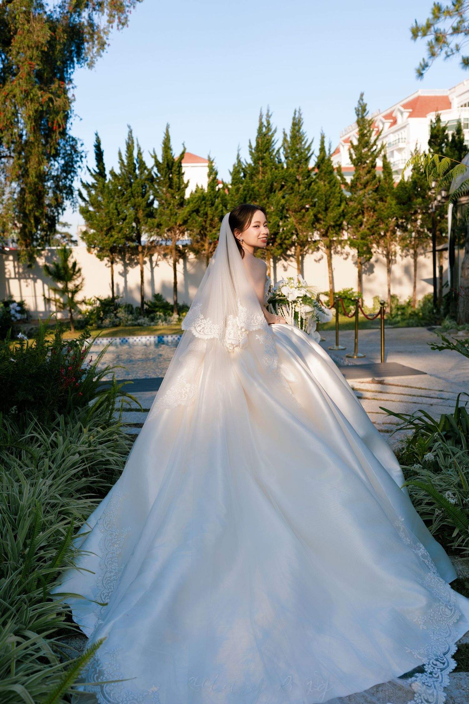 Bride in elegant white ball gown with long veil poses gracefully in a lush garden setting on her wedding day.