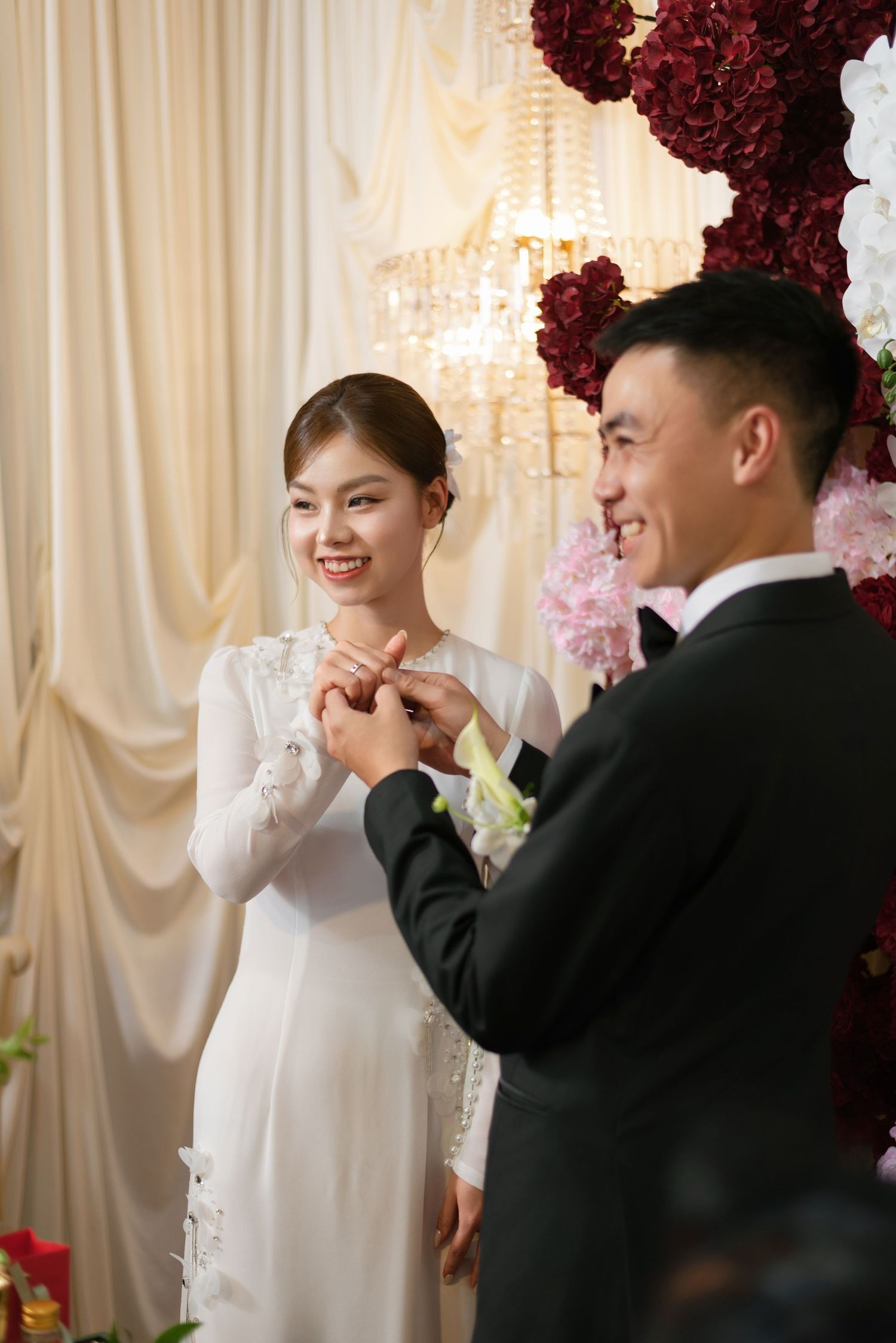 Smiling bride in white ao dai receives ring from groom in black suit at elegant wedding ceremony with floral backdrop.