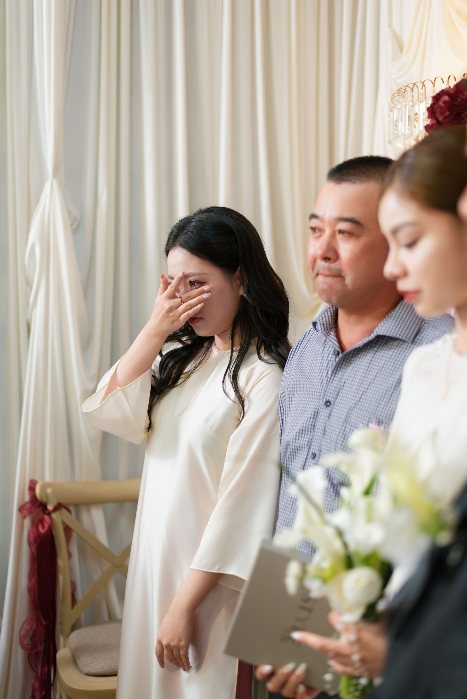 Emotional woman in white dress wiping tears at a wedding ceremony, standing beside a man and woman holding flowers.