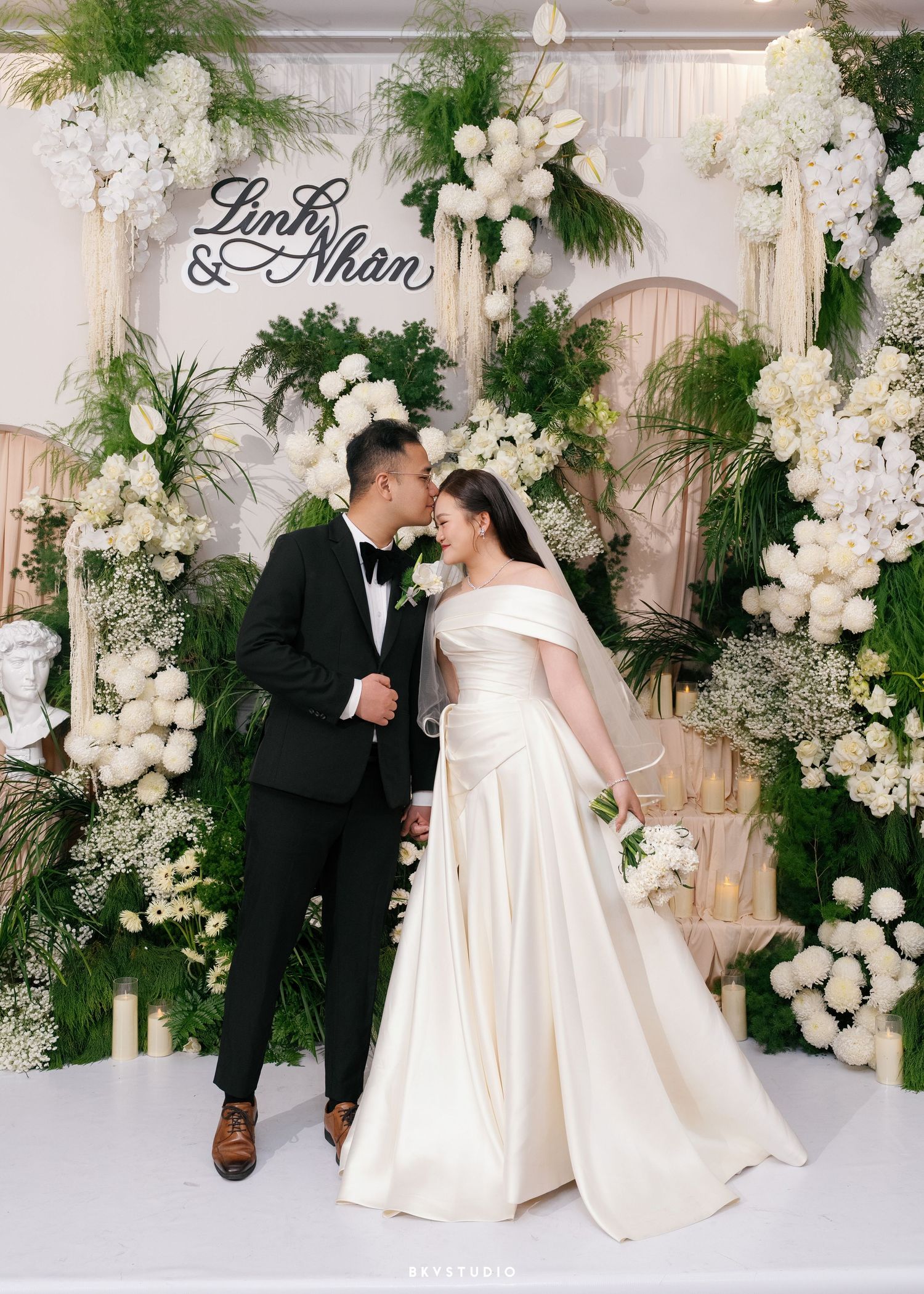 Couple Linh and Nhan share a romantic moment at their elegant wedding, surrounded by lush greenery and white floral arrangements.