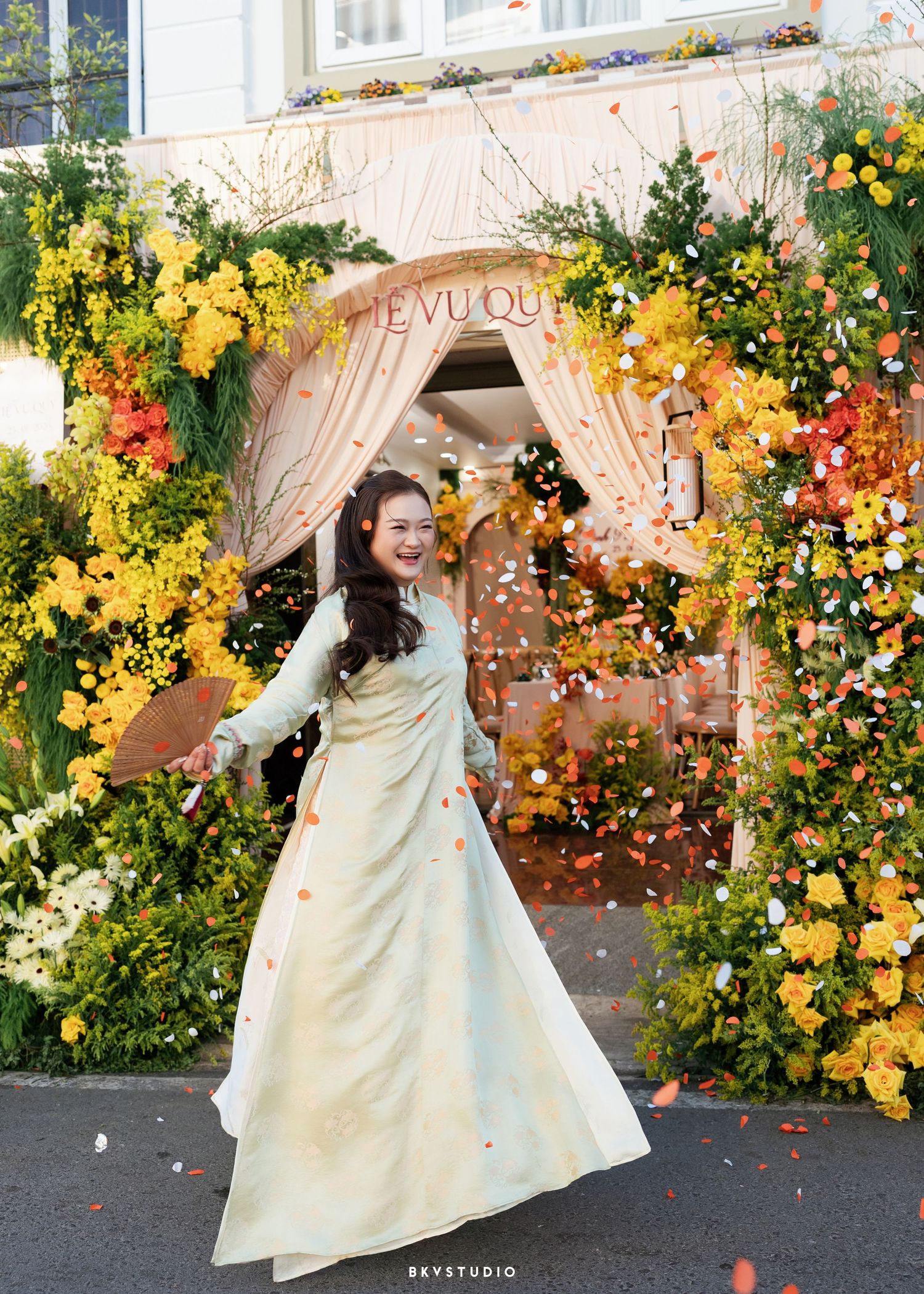 A joyful woman in a light green ao dai twirls under a floral arch adorned with yellow and orange blooms at a festive event.