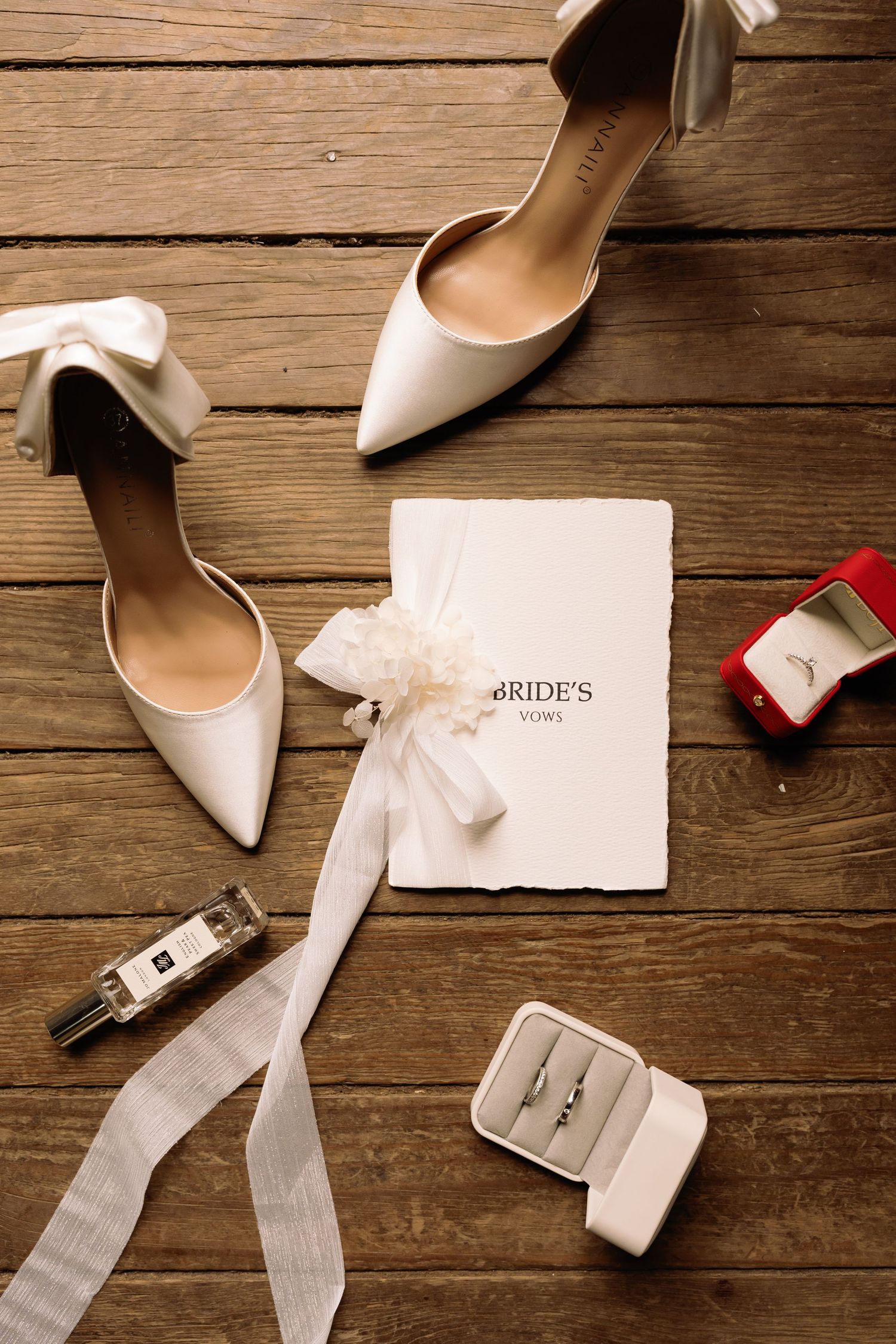 Elegant bridal flat lay with white heels, vow book, perfume, and ring boxes on rustic wood floor.