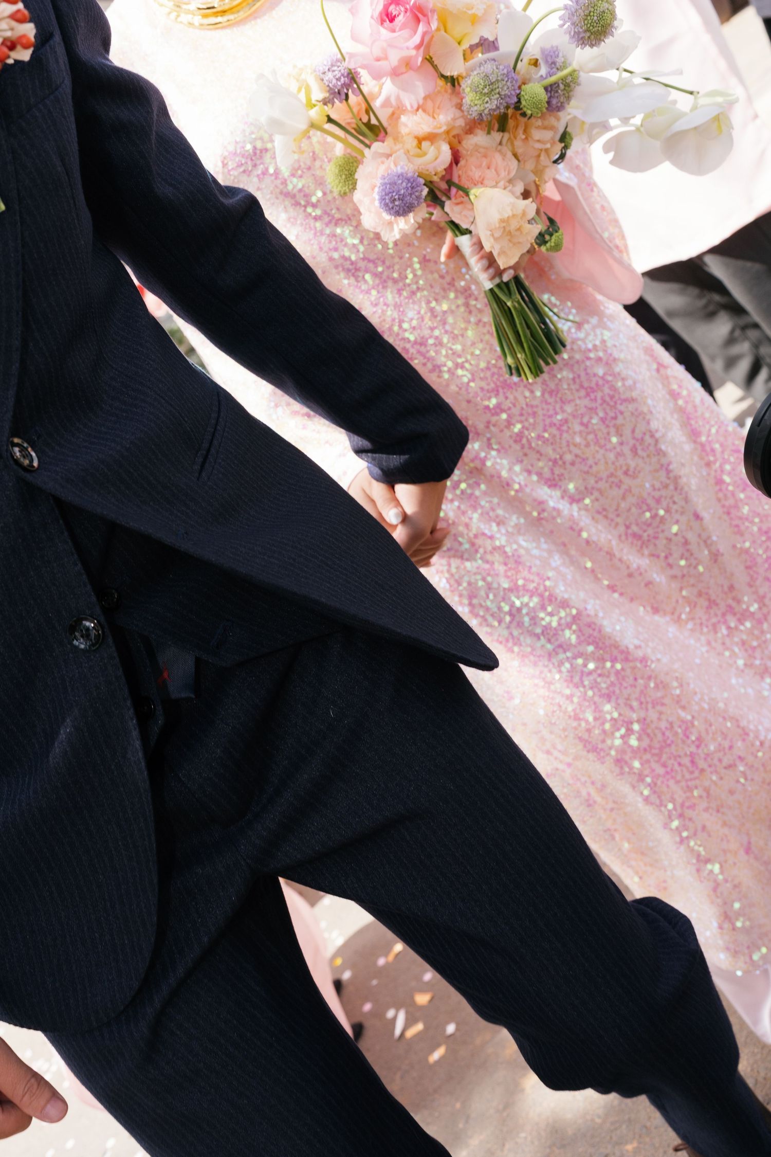 Groom in black suit holding bride's hand near pink sequin table with pastel wedding bouquet.