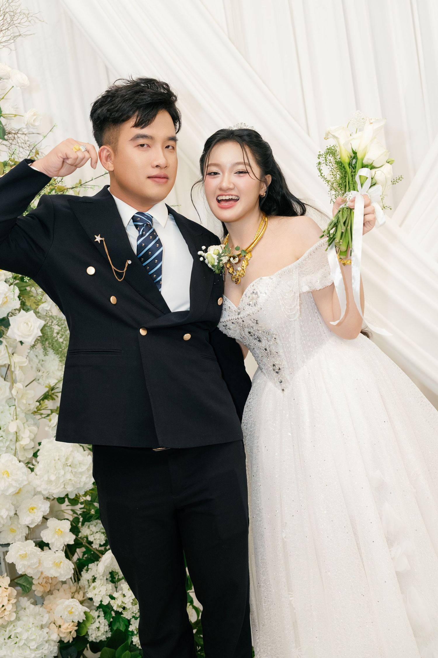 Laughing Vietnamese bride in white gown with gold jewelry and groom in black suit at floral wedding backdrop.