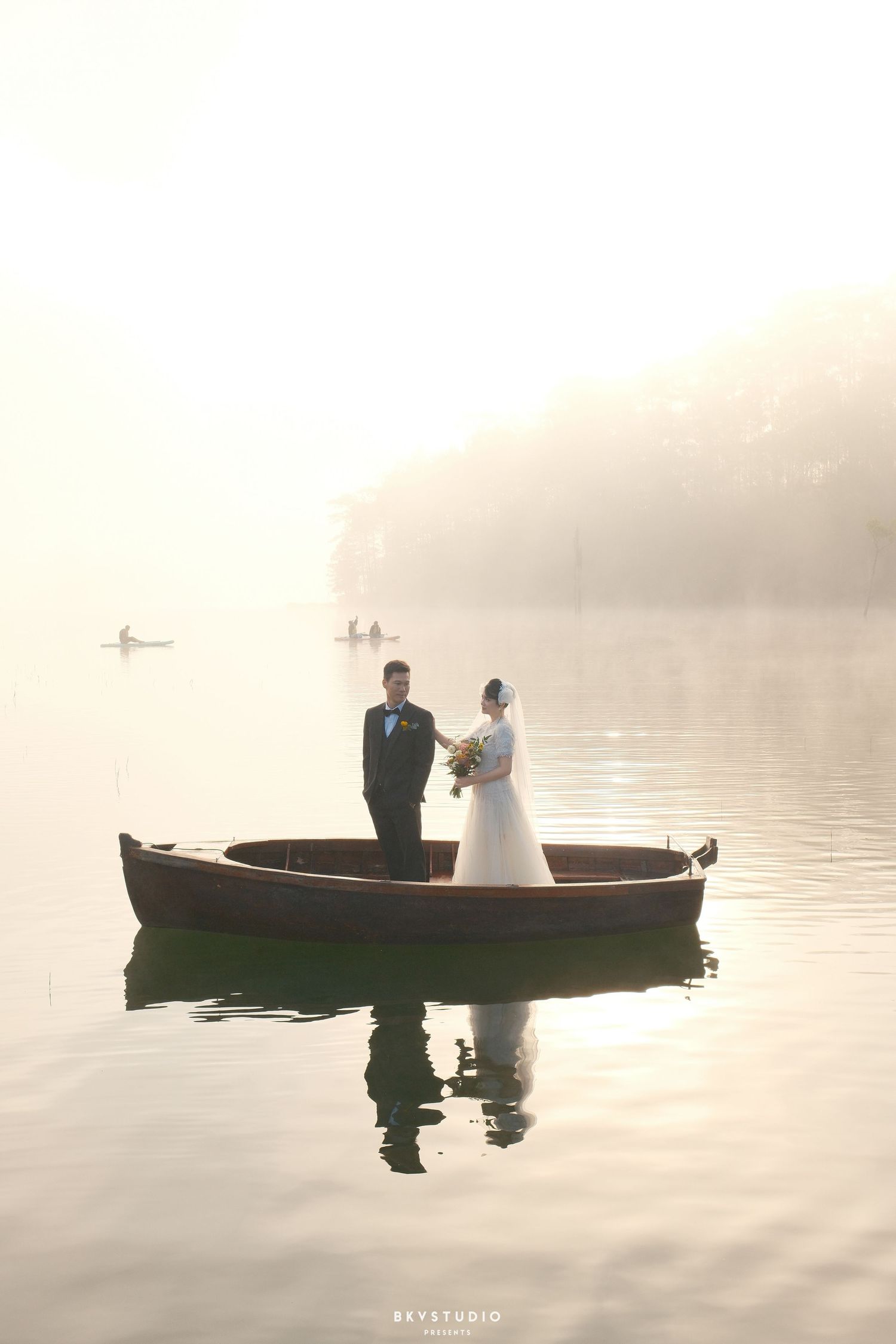 Bride and groom stand in wooden rowboat on calm misty lake, bride holding floral bouquet.