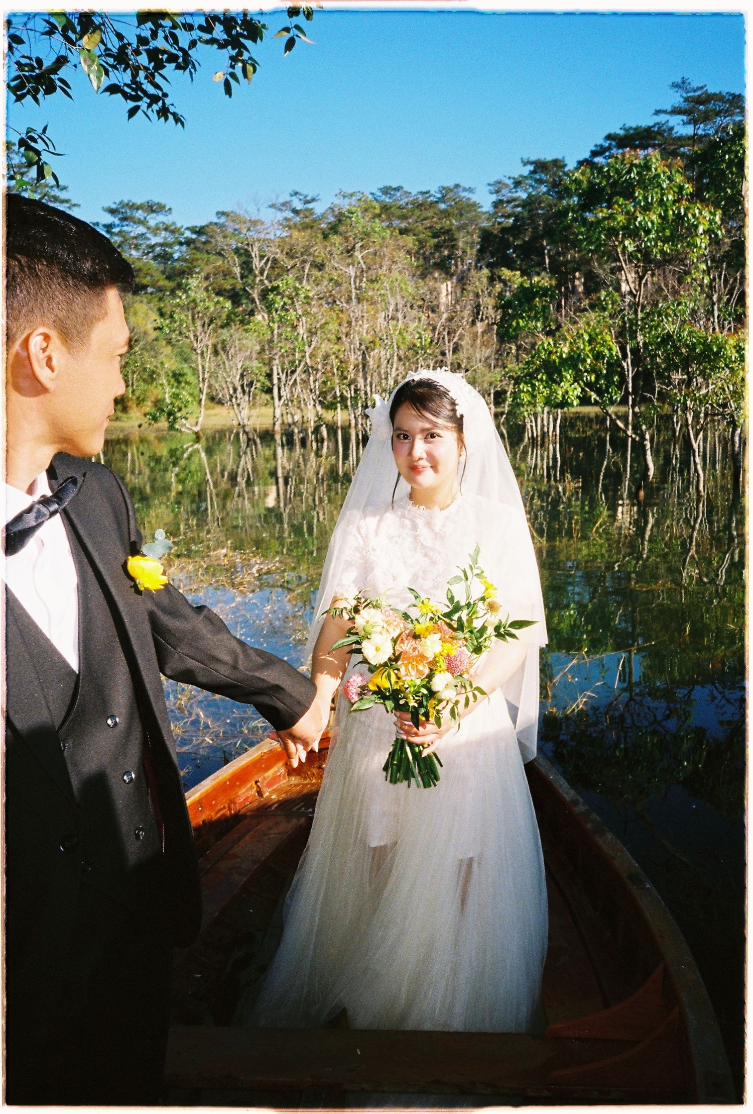 Bride and groom hold hands in wooden boat on reflective lake surrounded by lush green forest.