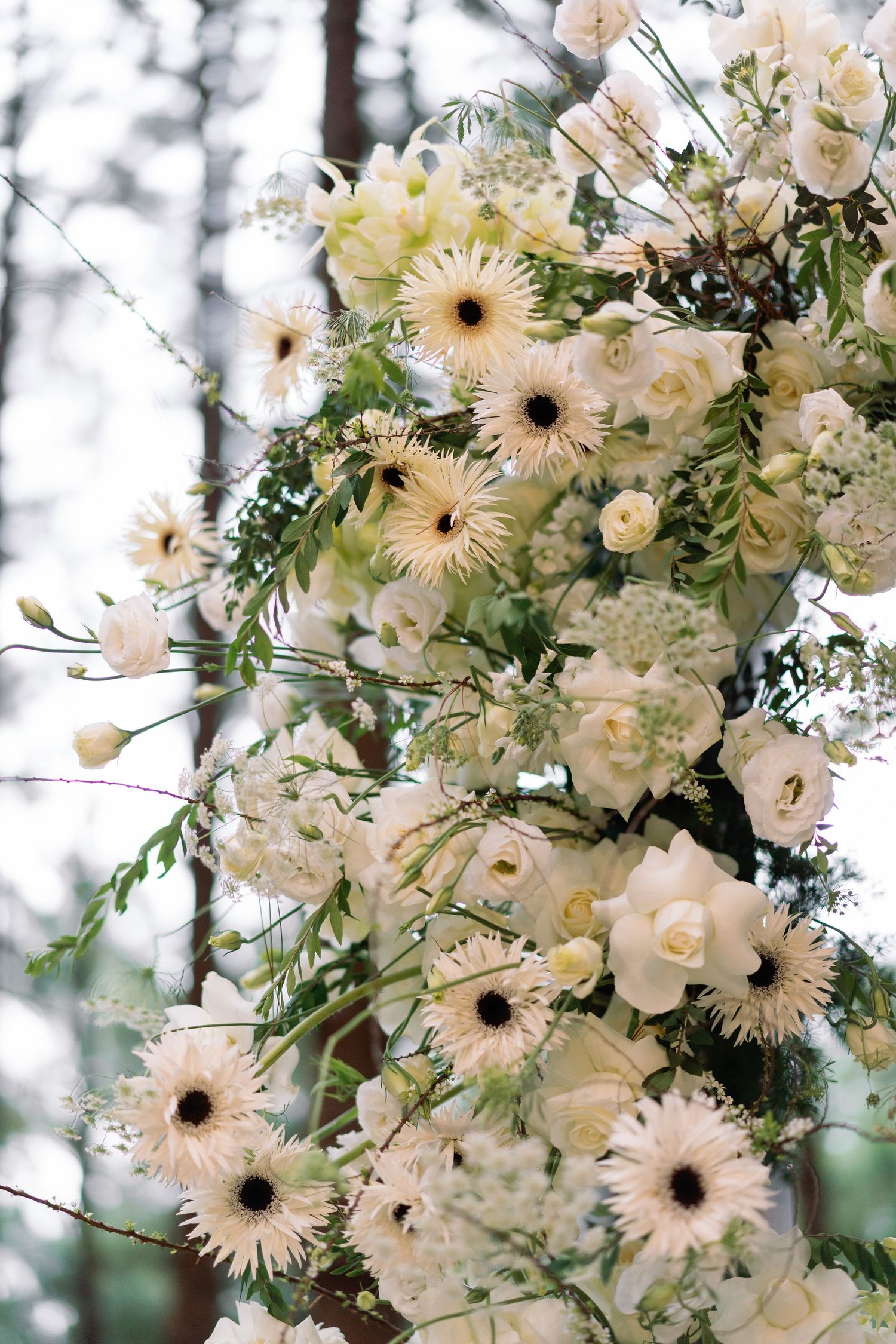 Elegant white floral wedding arch with roses, gerbera daisies, and greenery in a forest setting.