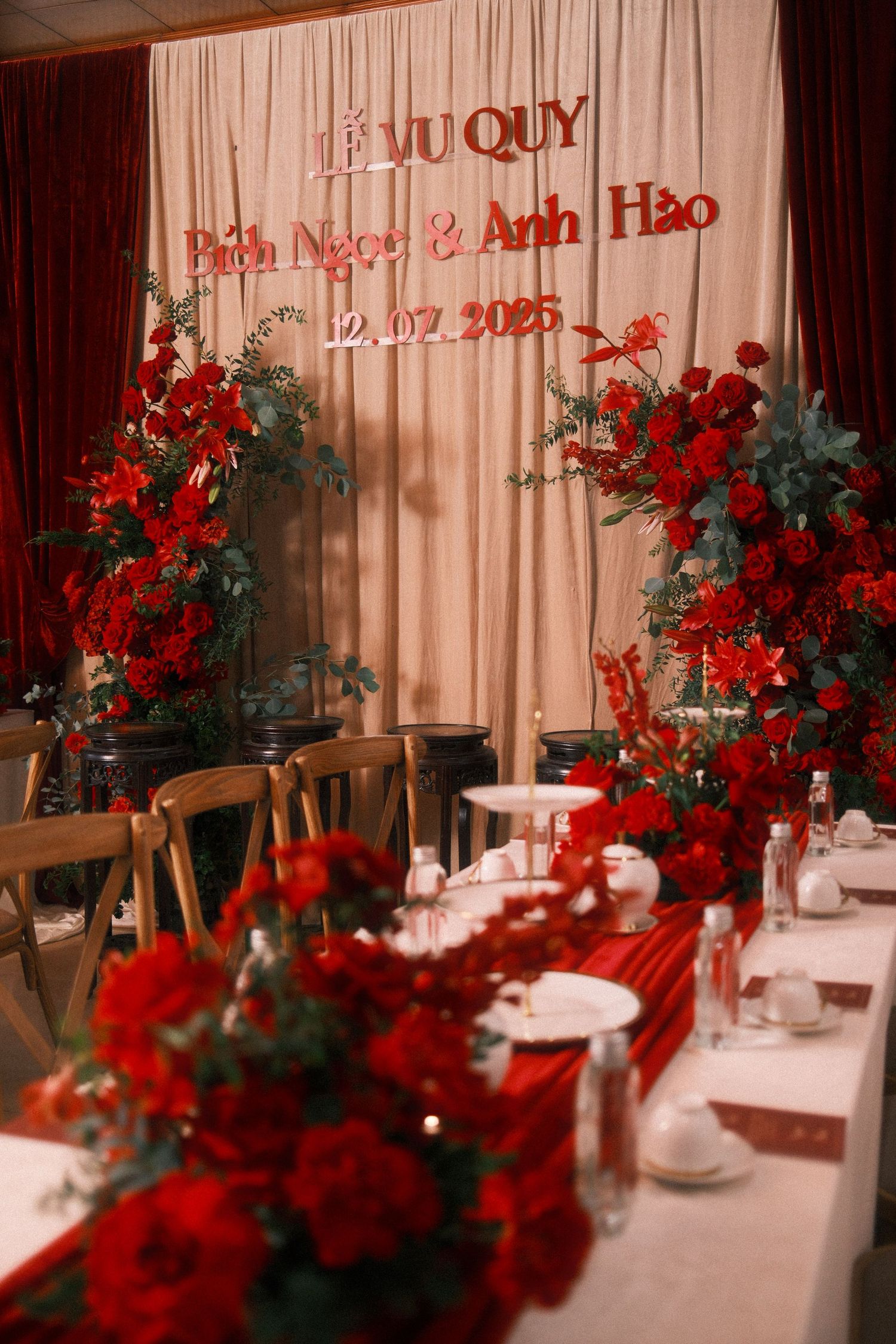 Vietnamese wedding reception decor with red roses, elegant table settings, and a backdrop reading Le Vu Quy dated 12.07.2025.