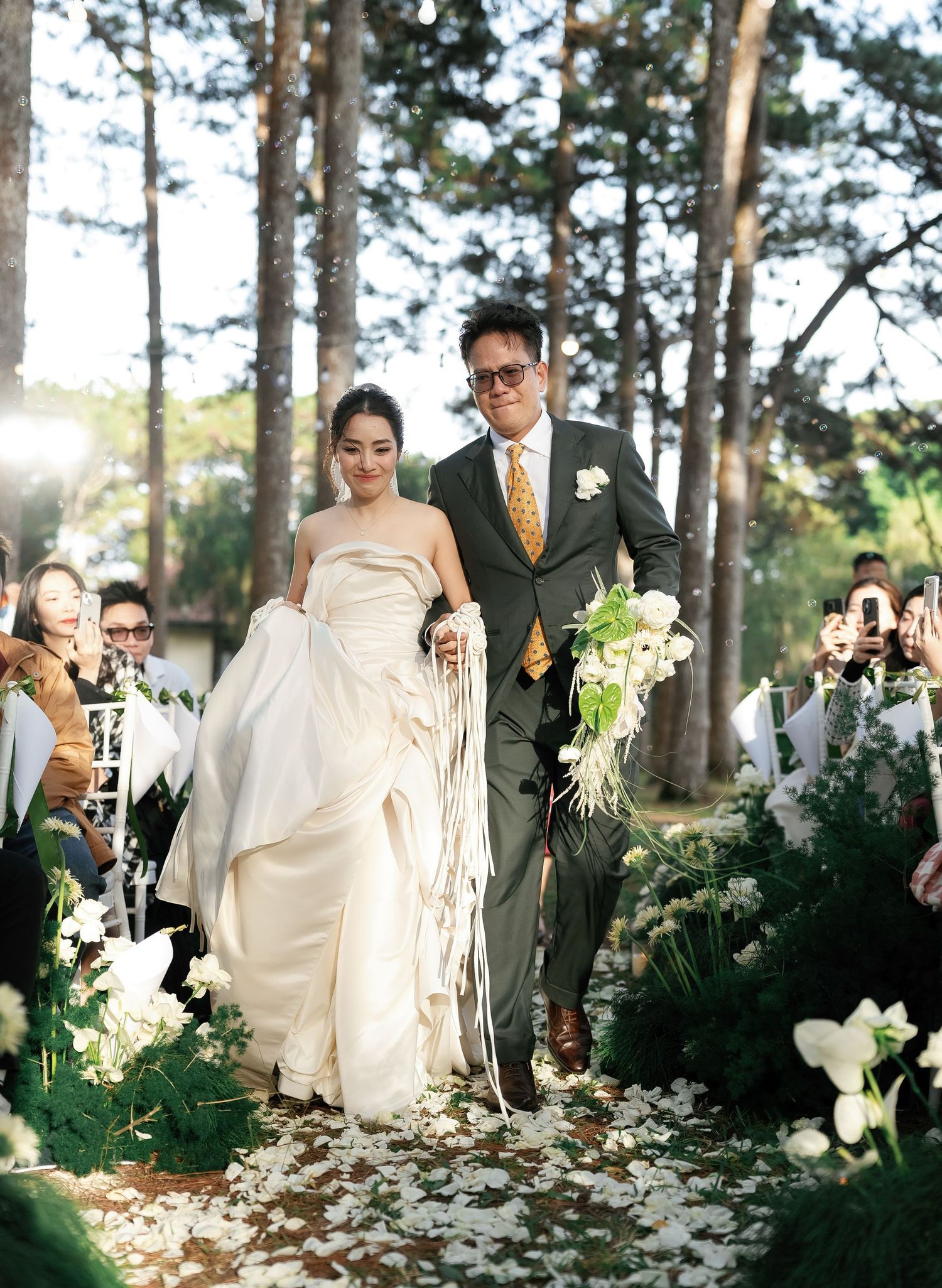 Happy newlywed couple walking down floral aisle after outdoor forest wedding ceremony, bride in white gown, groom in grey suit.