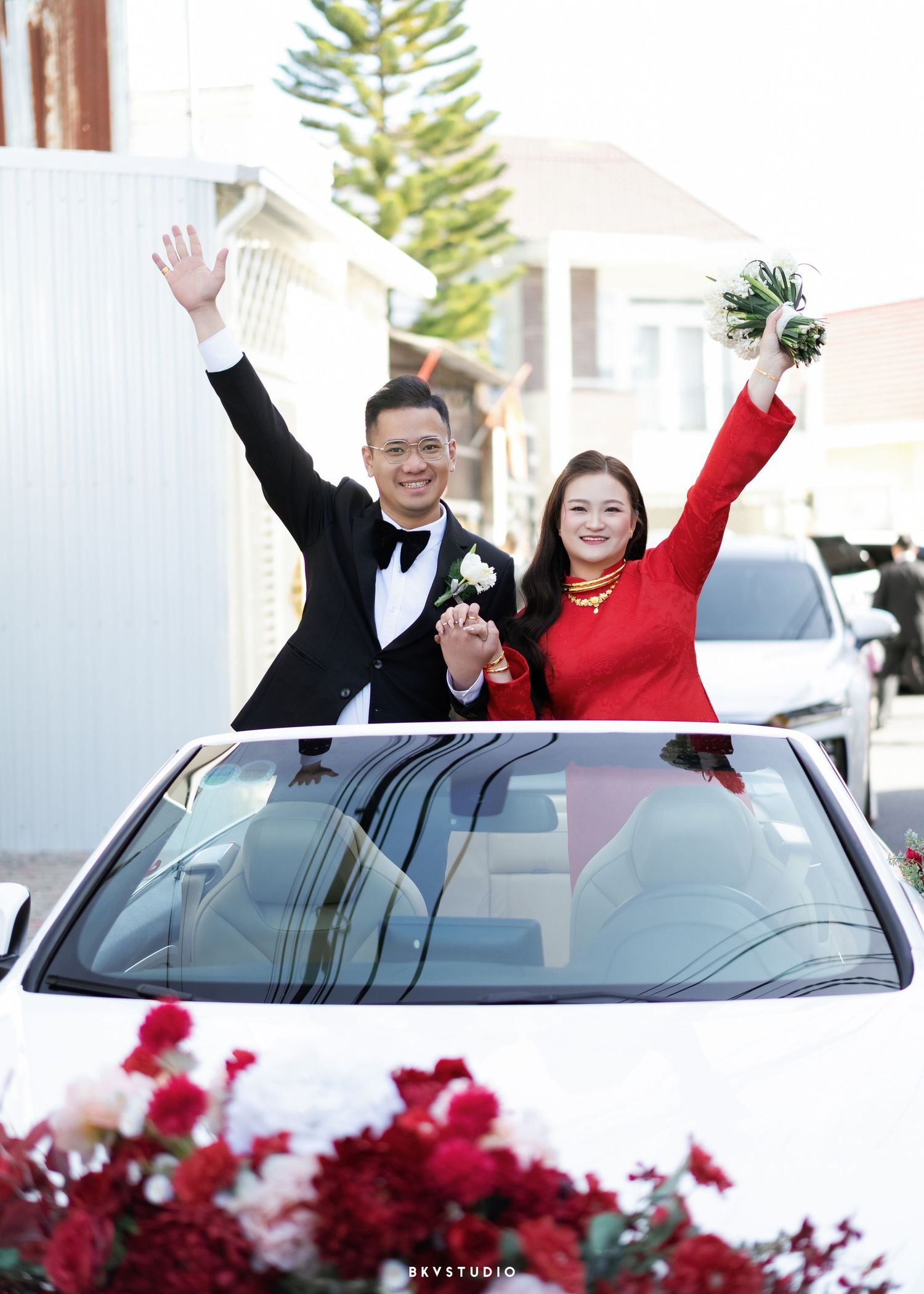 Happy couple in wedding attire waving from sunroof of white car decorated with red flowers.