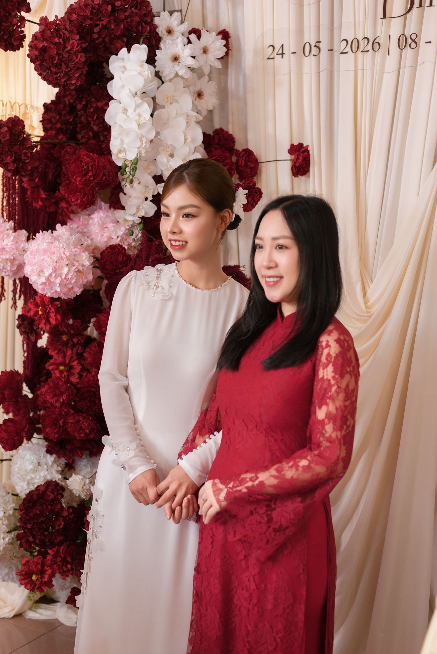 Two women in elegant ao dai dresses pose together at a Vietnamese wedding ceremony with floral backdrop.