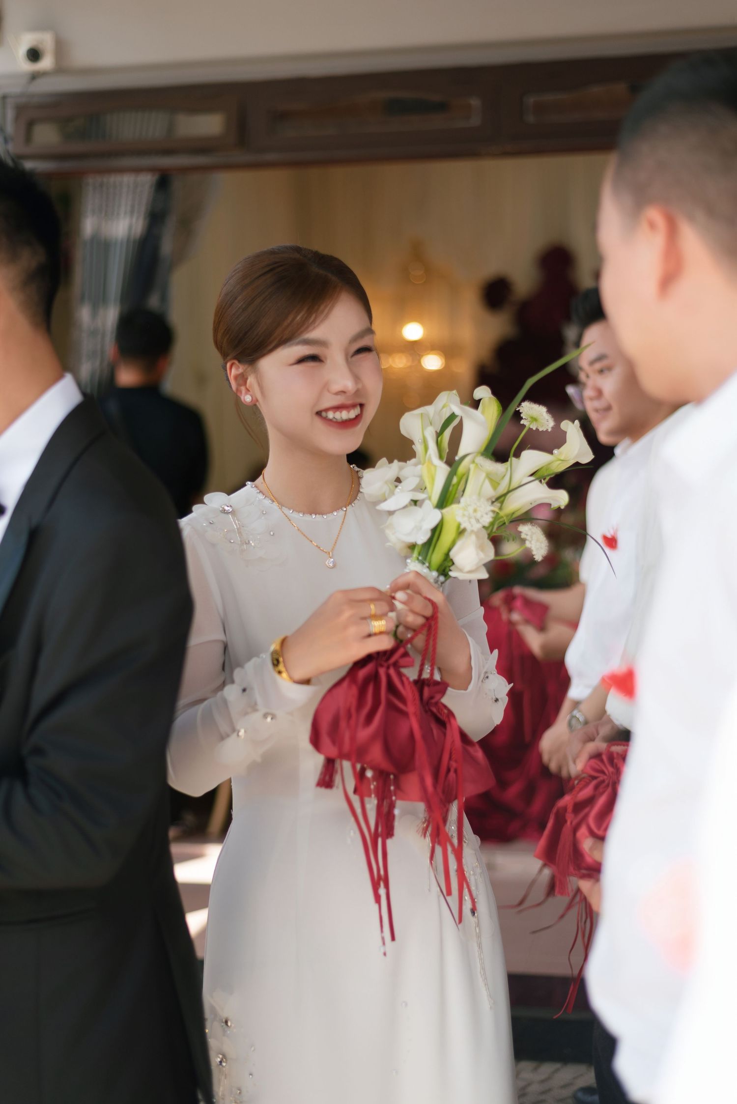 Smiling bride in white dress holds calla lilies and red accessories at an elegant wedding ceremony.