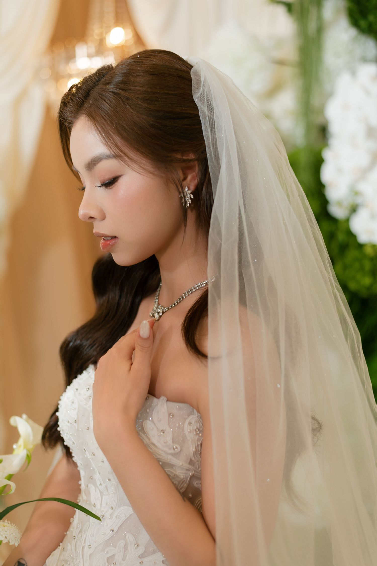 A bride in a white lace gown and veil touches her diamond necklace, eyes downcast, surrounded by white flowers.