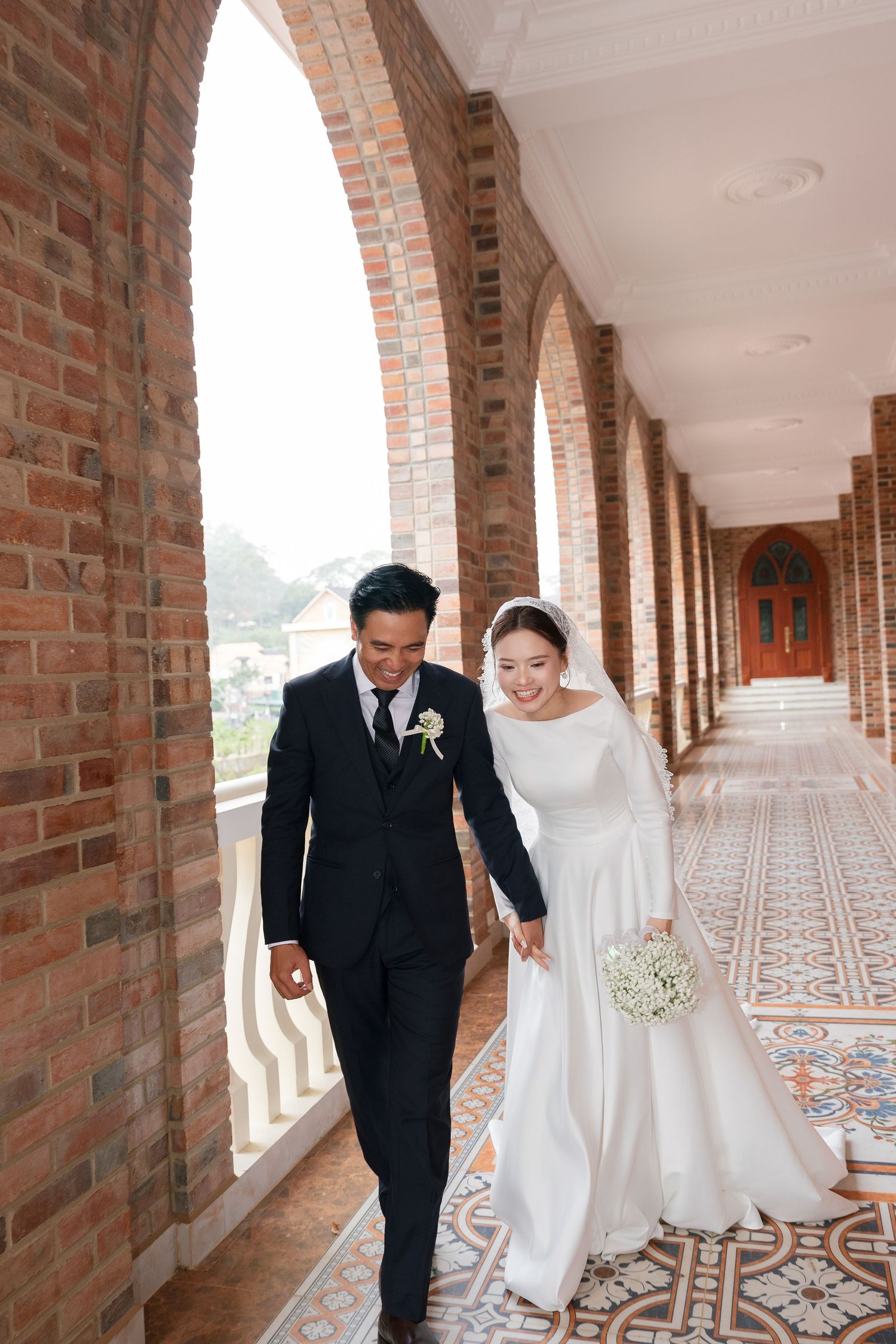 A smiling bride and groom walk hand-in-hand through an elegant brick archway corridor on their wedding day.