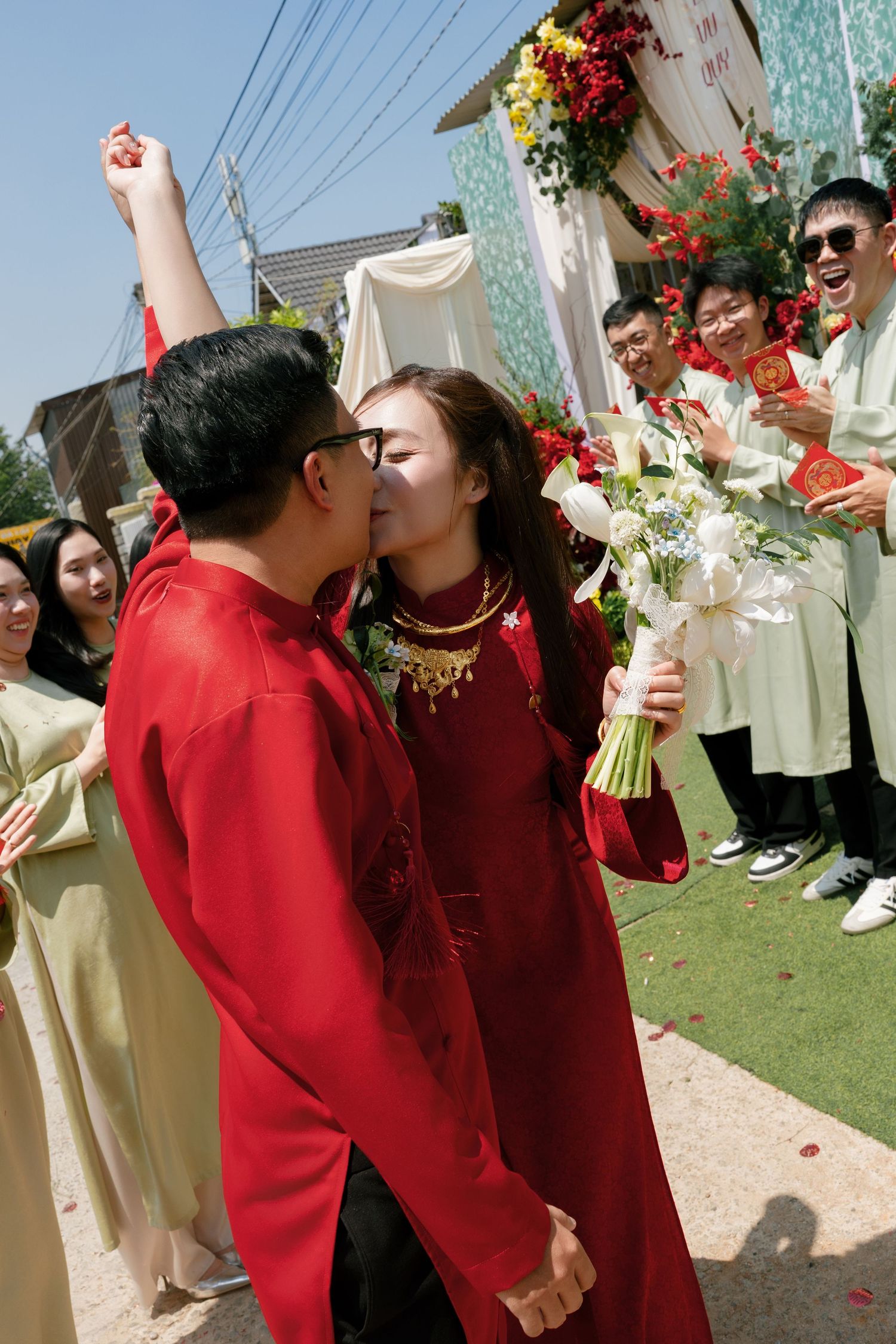 Vietnamese couple in red ao dai kissing at traditional wedding ceremony, surrounded by cheering guests holding flowers.