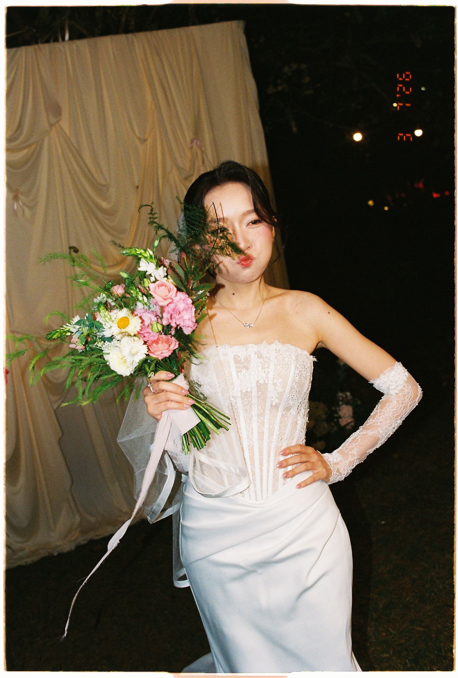 Woman in white corset gown holding pink and white floral bouquet at night, film photography aesthetic.