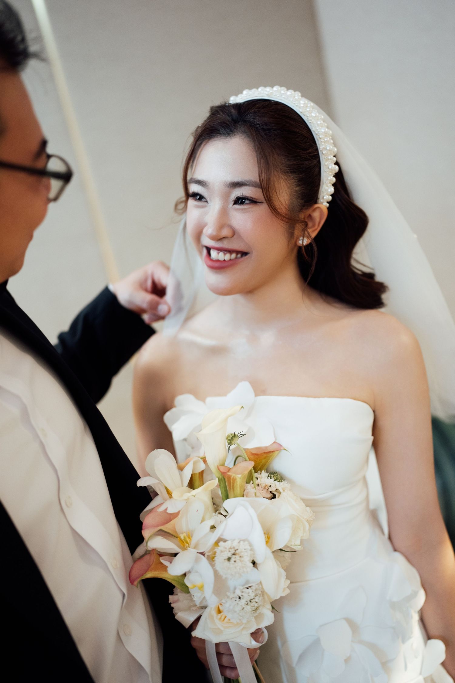 Smiling bride in white strapless gown holding white lily bouquet with pearl headband and veil at wedding ceremony.