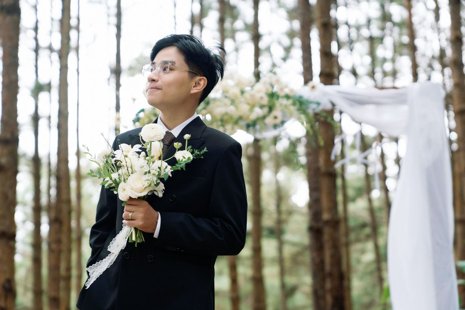 Groom in black suit holding white floral bouquet at outdoor forest wedding ceremony.