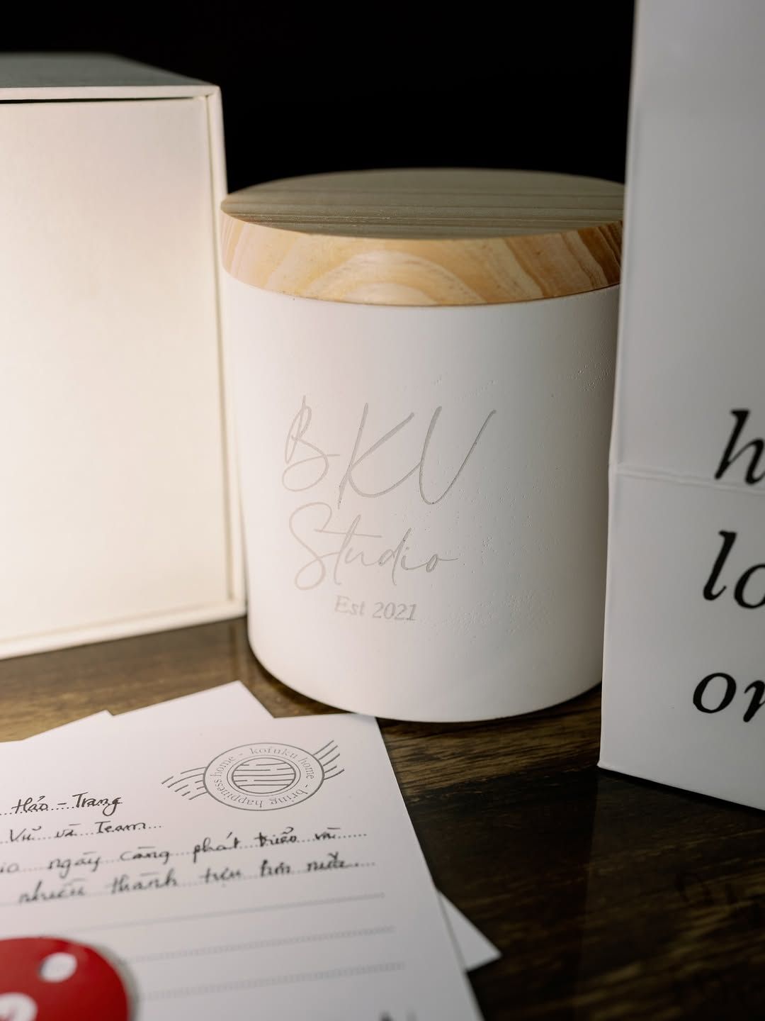 BKV Studio Est 2022 white candle with wooden lid, handwritten note, and gift box on wooden table.