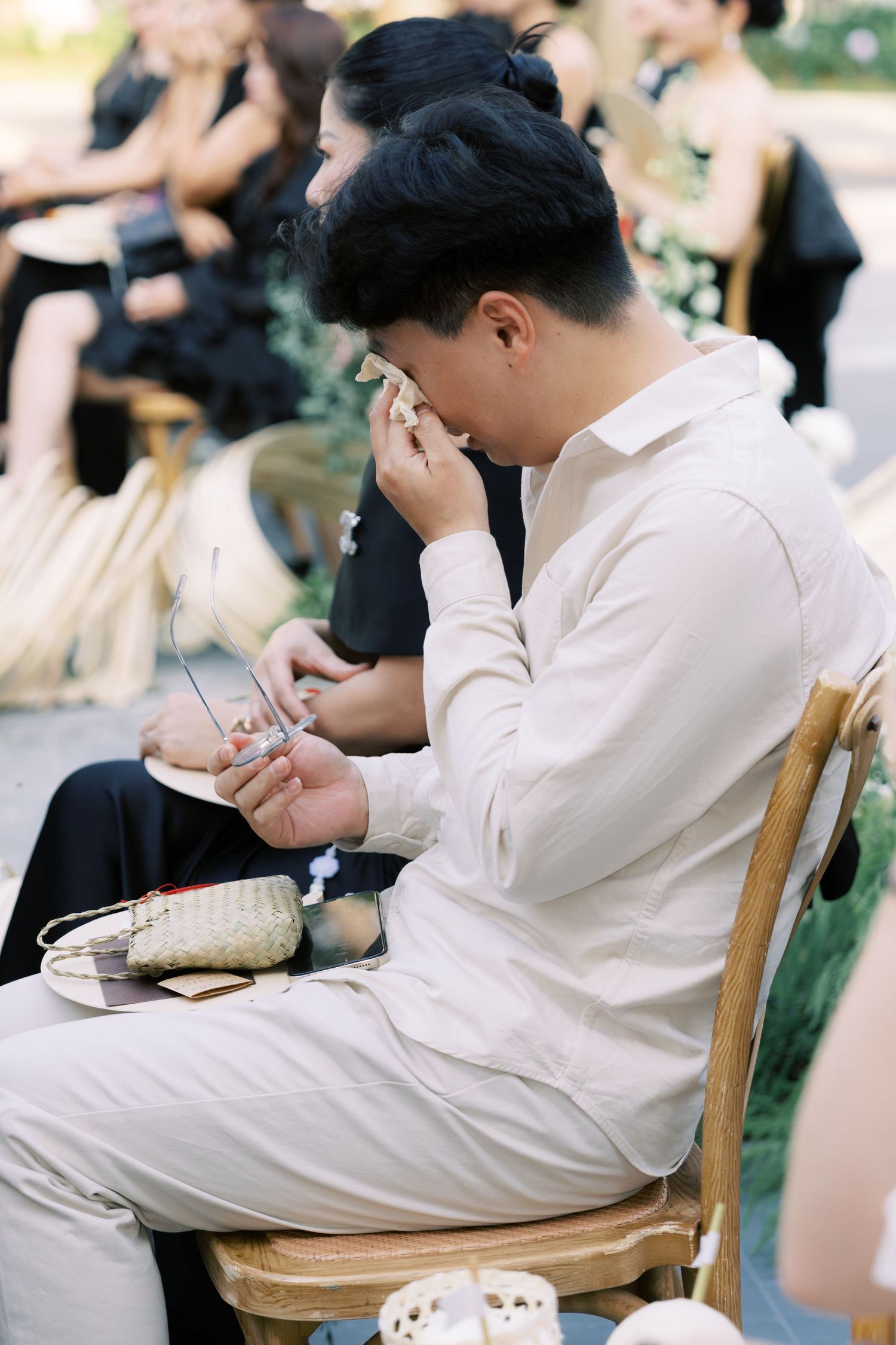 Emotional male wedding guest wiping tears while seated at an outdoor wedding ceremony.