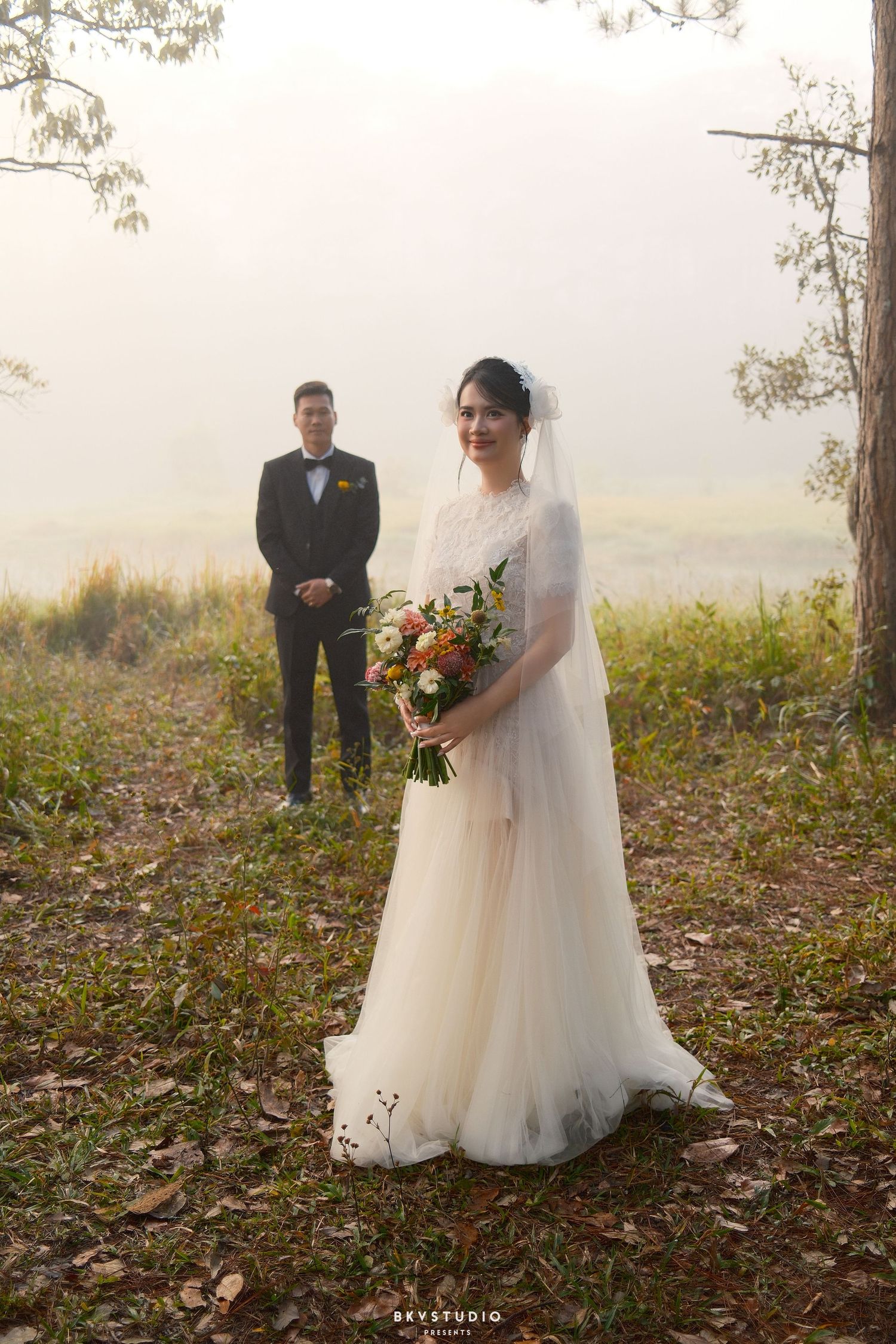 A bride in a white gown holds a bouquet while the groom stands in the misty background outdoors.