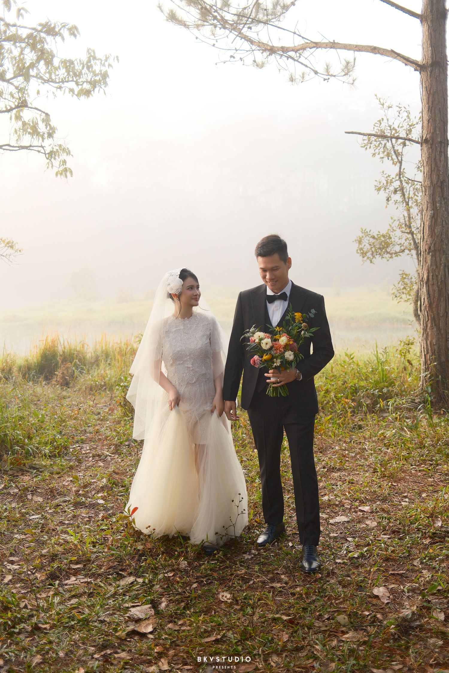 Wedding couple walks hand in hand through misty forest, groom in black tuxedo, bride in white gown.