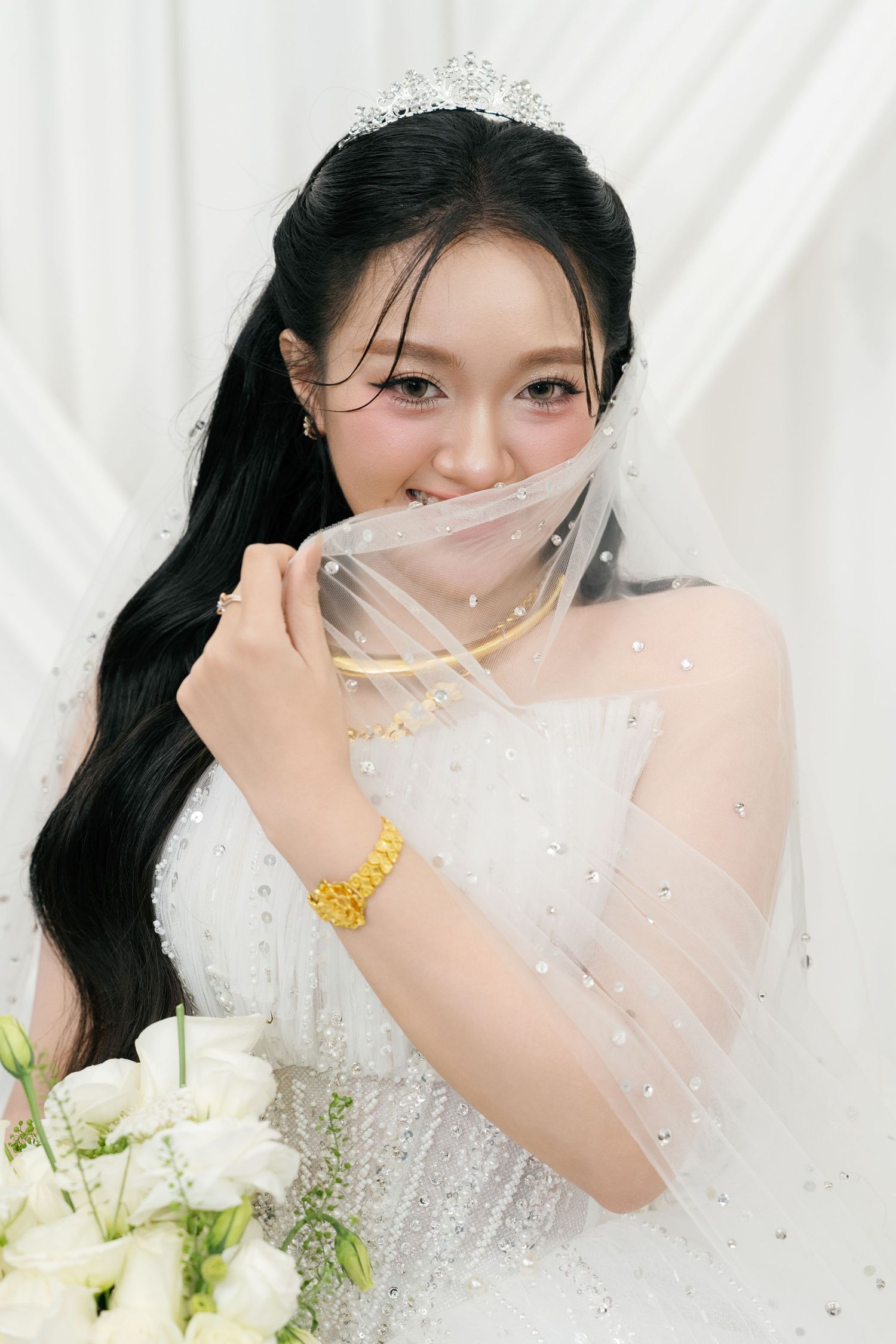 Beautiful Asian bride in white gown with tiara, veil, and gold jewelry holding white flower bouquet.