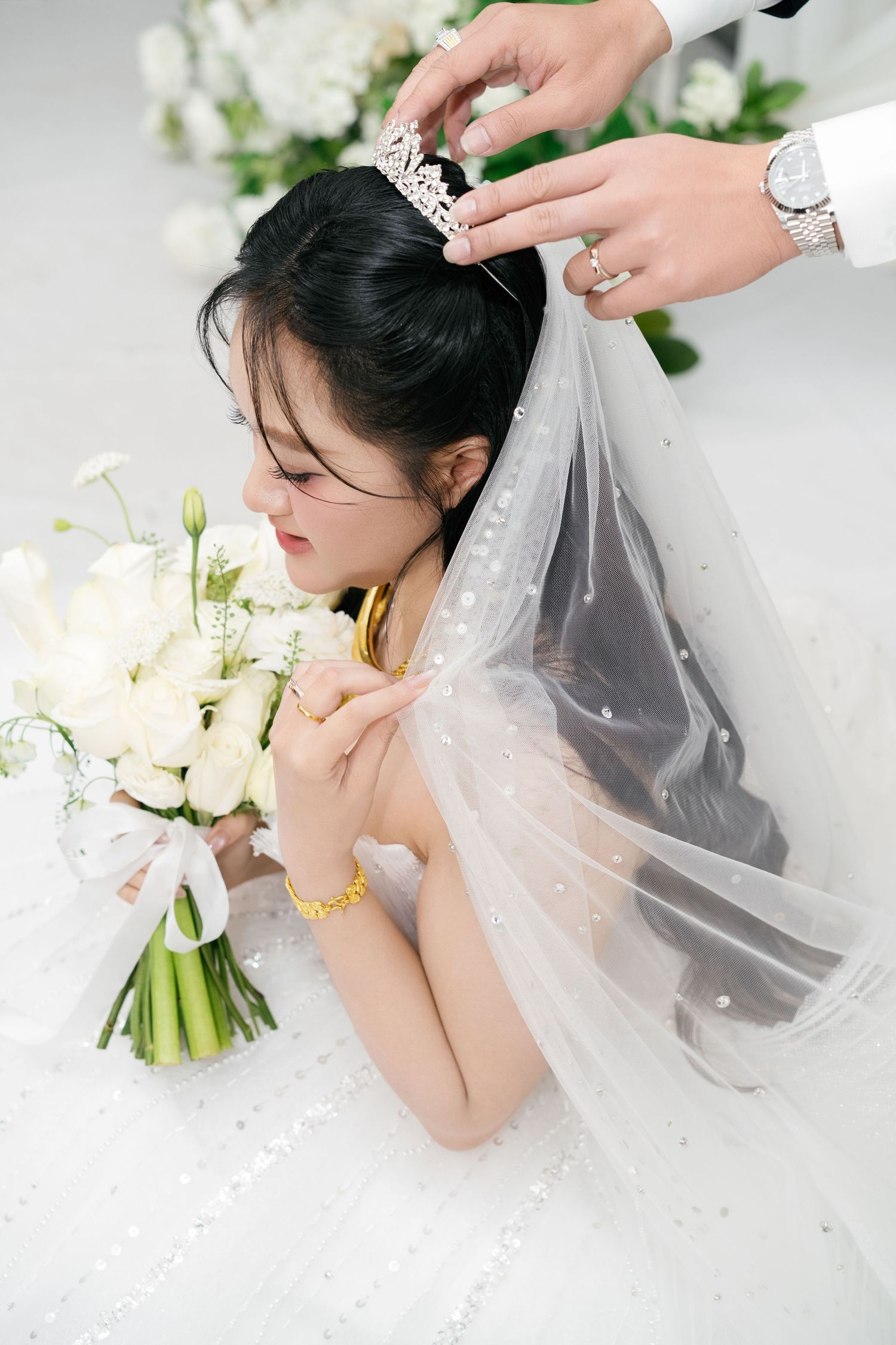 Groom places a diamond tiara on bride wearing a white wedding gown and veil while she holds flowers.