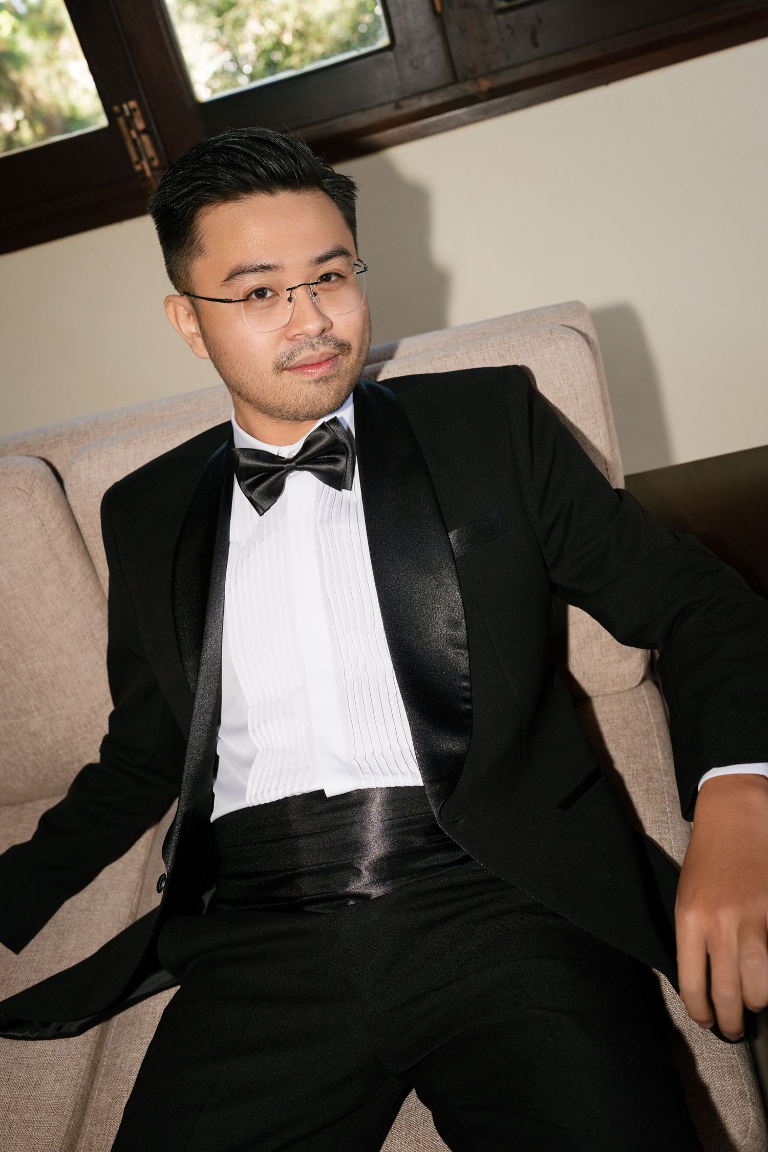 Handsome groom in black tuxedo and bow tie sitting relaxed on a beige armchair.