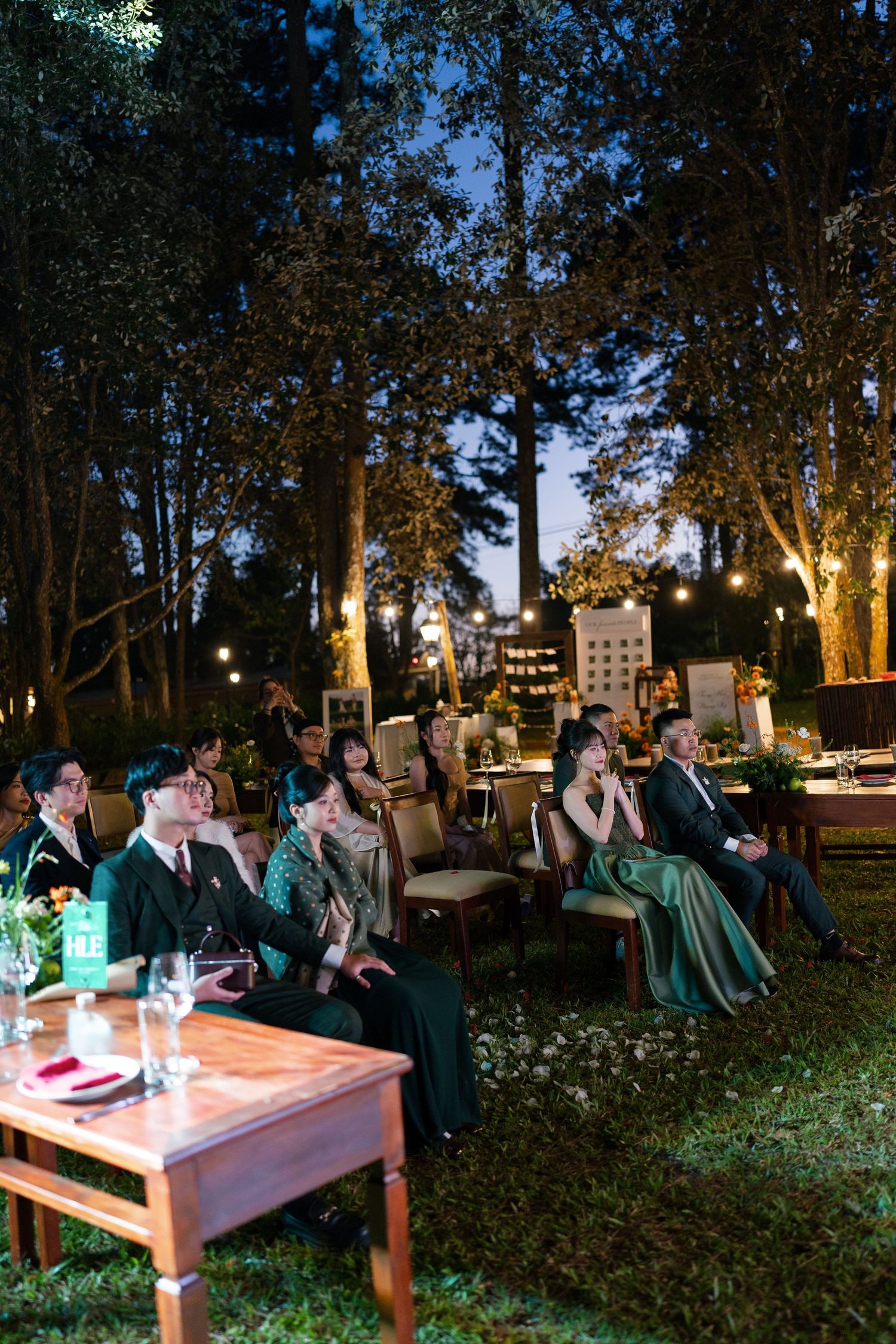 Wedding guests seated outdoors at night under trees with warm string lights during garden ceremony.