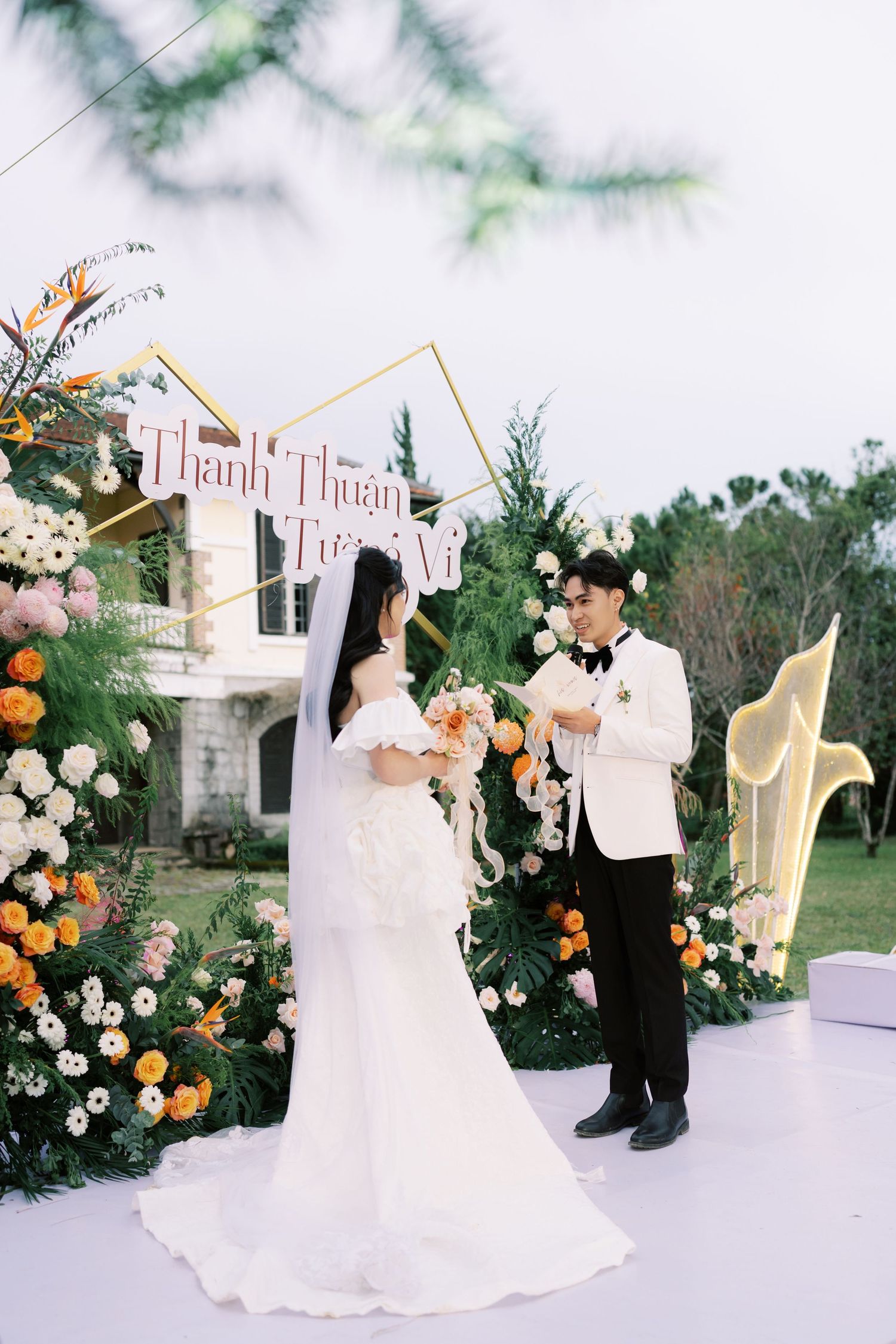 Bride and groom exchange vows at an outdoor wedding ceremony with floral arch and Vietnamese signage.
