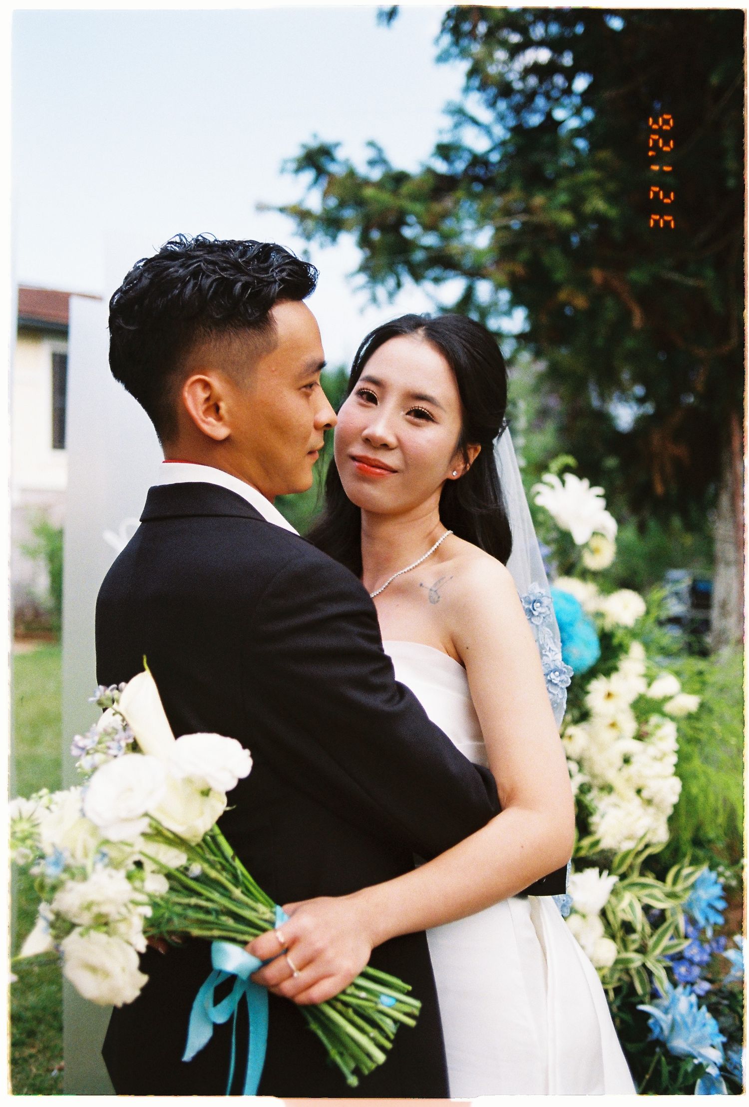 Asian couple in wedding attire embracing outdoors, bride holding white and blue bouquet with teal ribbon.
