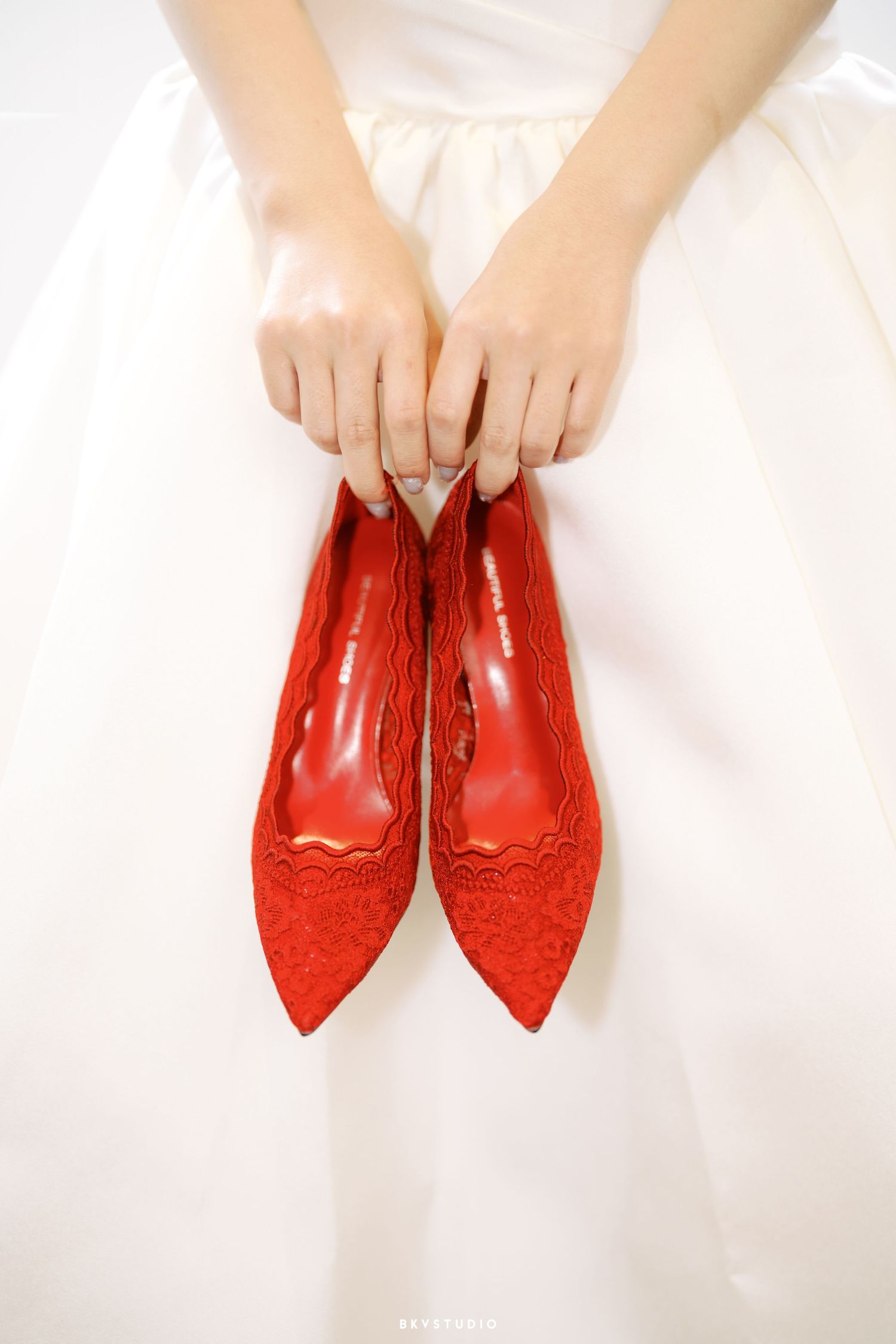 Bride in white dress holding elegant red lace pointed-toe flats for wedding day.