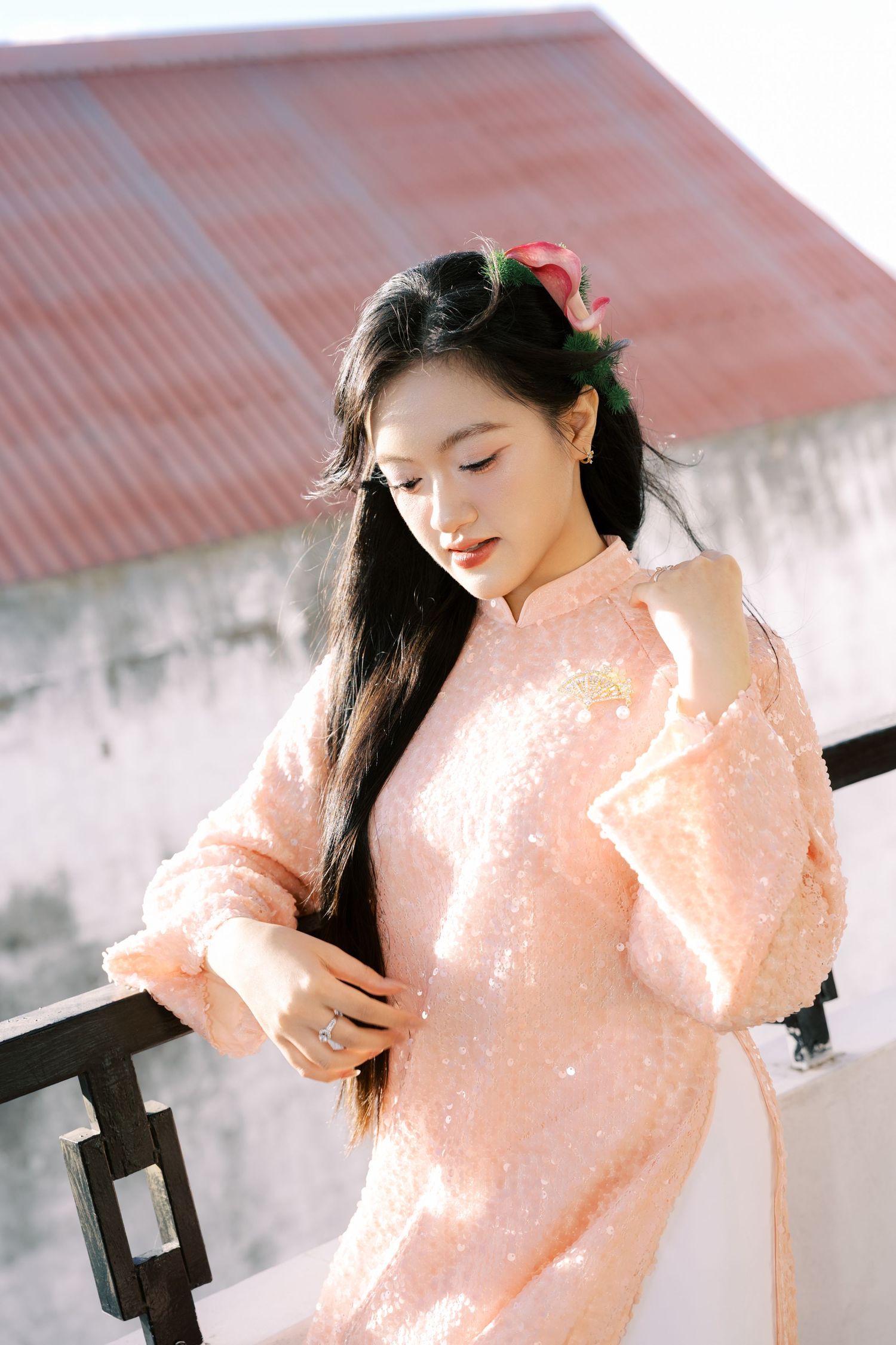Young Vietnamese woman in pink sequined ao dai traditional dress posing on sunlit balcony.