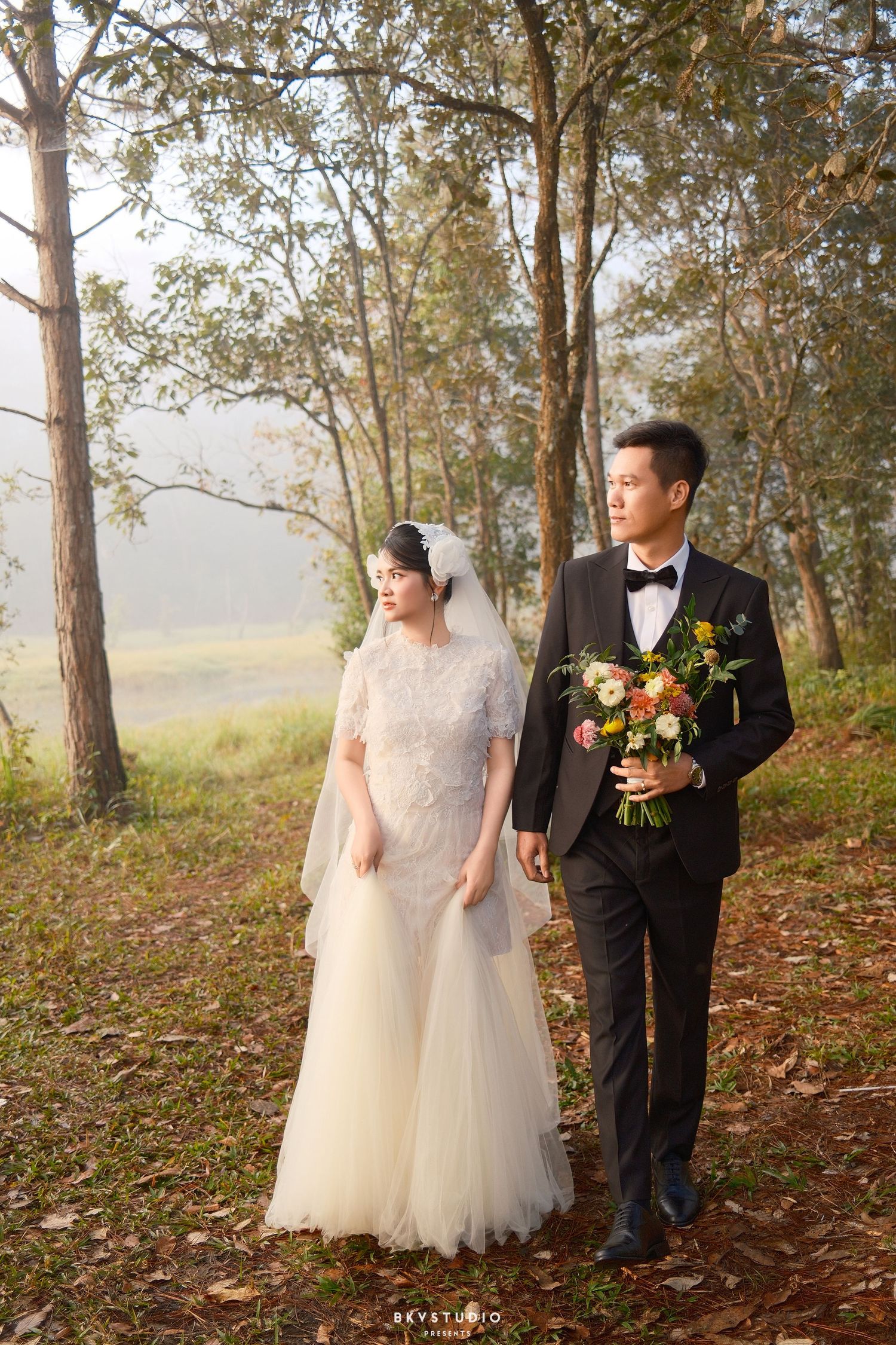 Wedding couple walks through forest, bride in lace gown with veil, groom in black suit holding colorful bouquet.